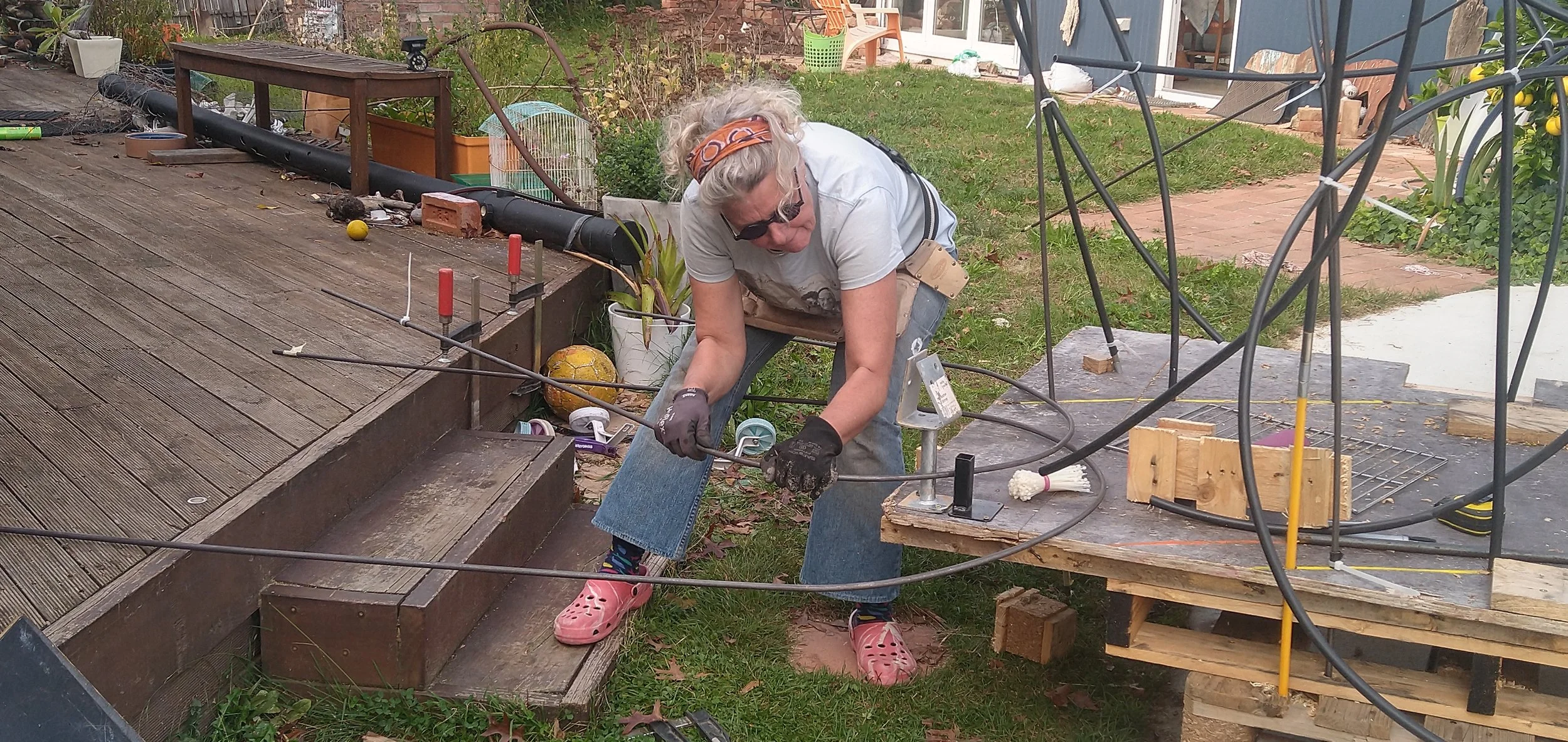 Woman working on a a sculpture project outdoors, using tools and wearing gloves, bending mild steel into shape, with wooden stairs and garden tools around her.