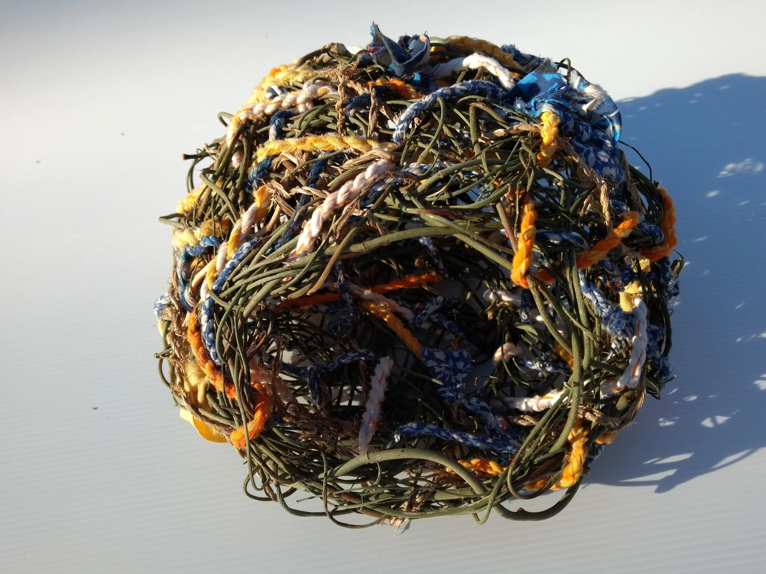 A spherical sculpture made of intertwined vines and colorful fabric, set against a white background.