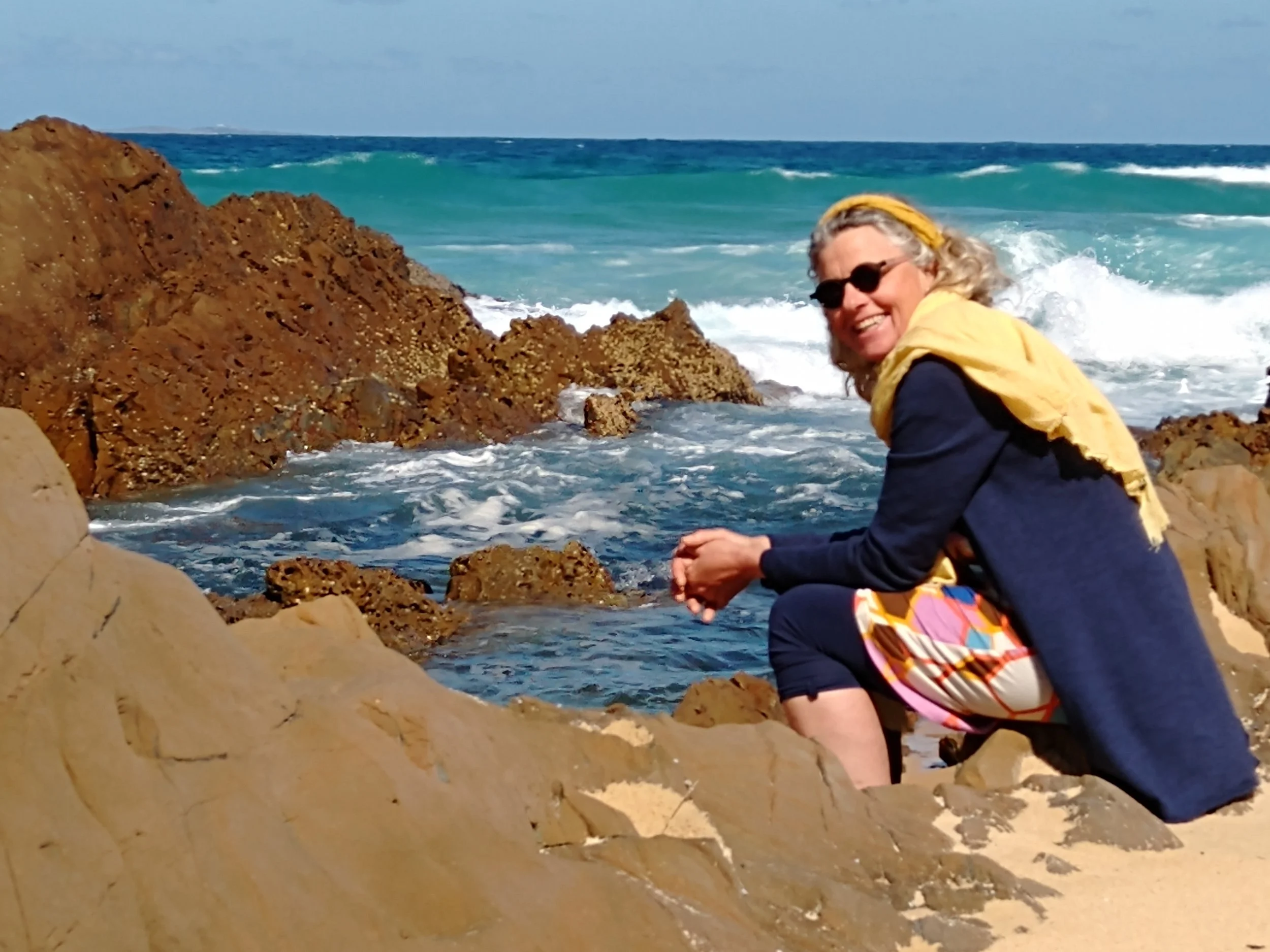 A woman sitting on rocks by the ocean, smiling, wearing sunglasses, a yellow scarf, a blue jacket, and patterned shorts, with waves and blue water in the background.