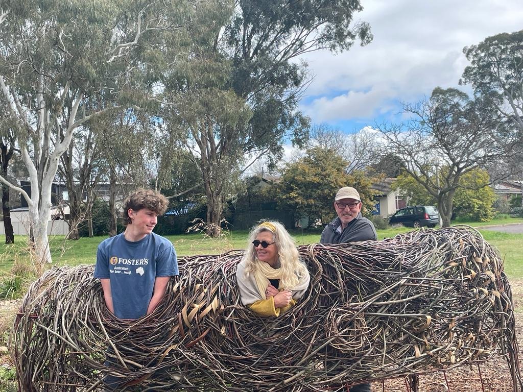 Three people sitting inside a large, circular sculpture made of intertwined sticks or branches in an outdoor park with trees and houses in the background.