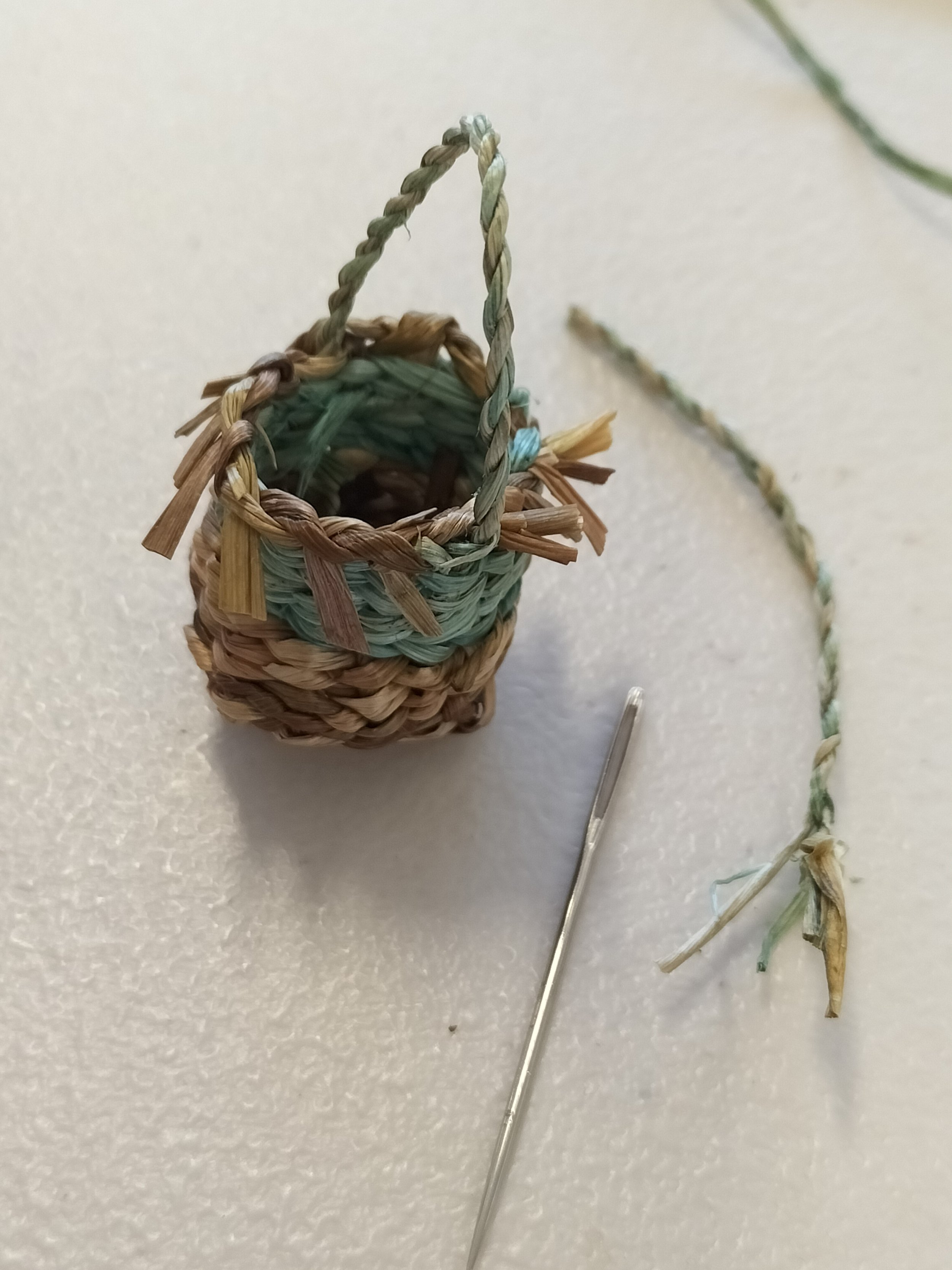 A finished miniature twined basket with a twisted rope handle, containing loose green and brown plant fibres, with some additional fibres and a needle lying on a white textured surface.