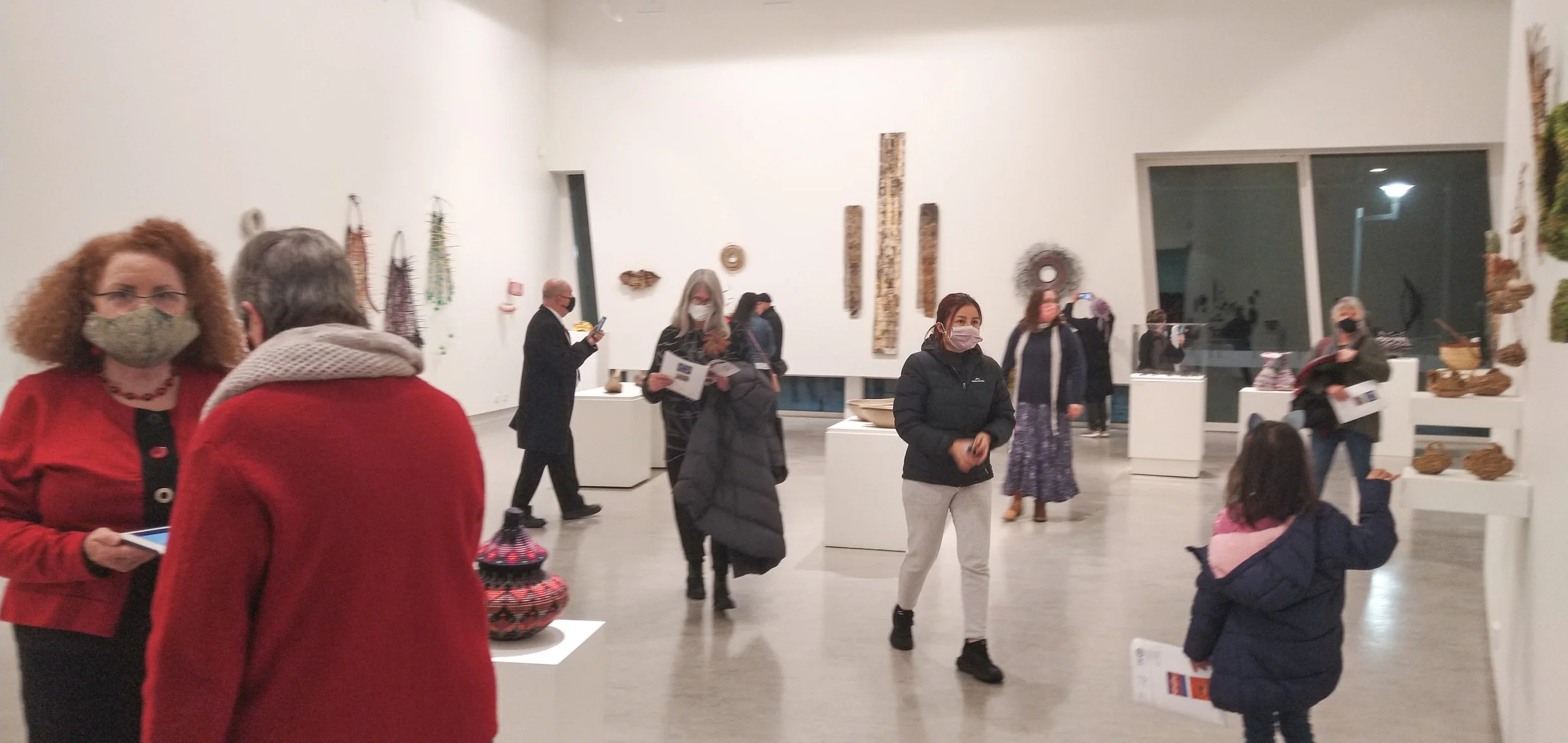 People wearing masks inside an art gallery, observing and discussing various artwork and sculptures displayed on white pedestals and walls.