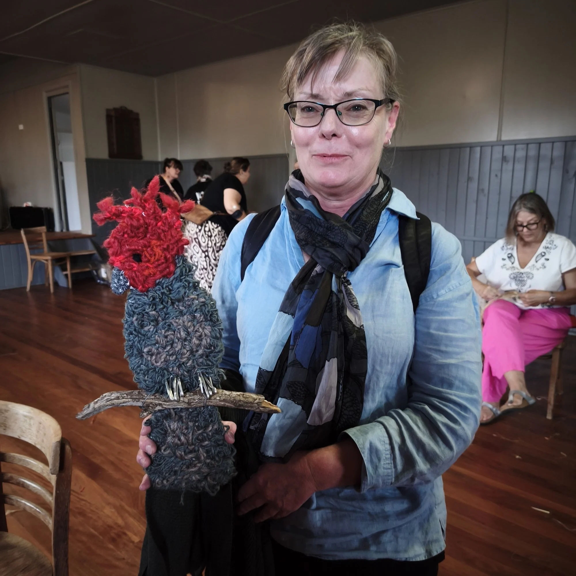 A woman with short blonde hair, glasses, and a black scarf is holding a colourful, fuzzy black bird sculpture. She is smiling slightly and standing inside a room with wooden floors and chairs, and there are other people in the background.