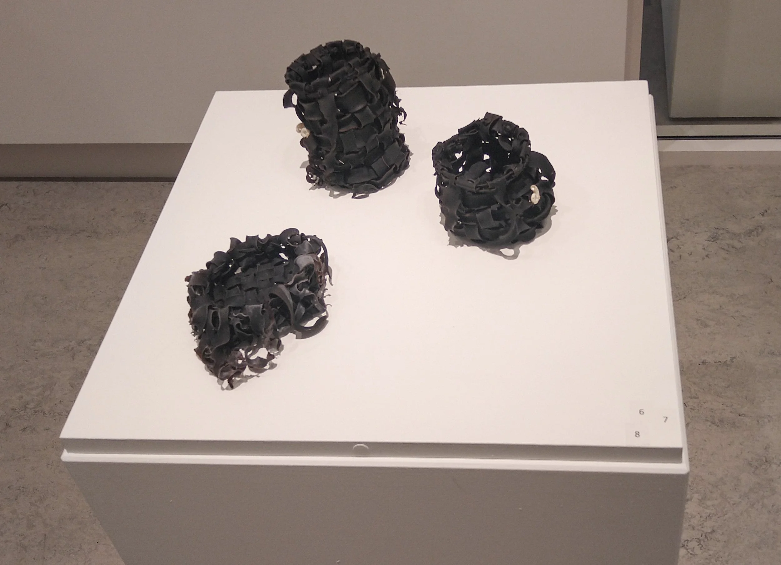 Three dark, ribbon-like sculptures with pearl accents displayed on a white pedestal in an art gallery.
