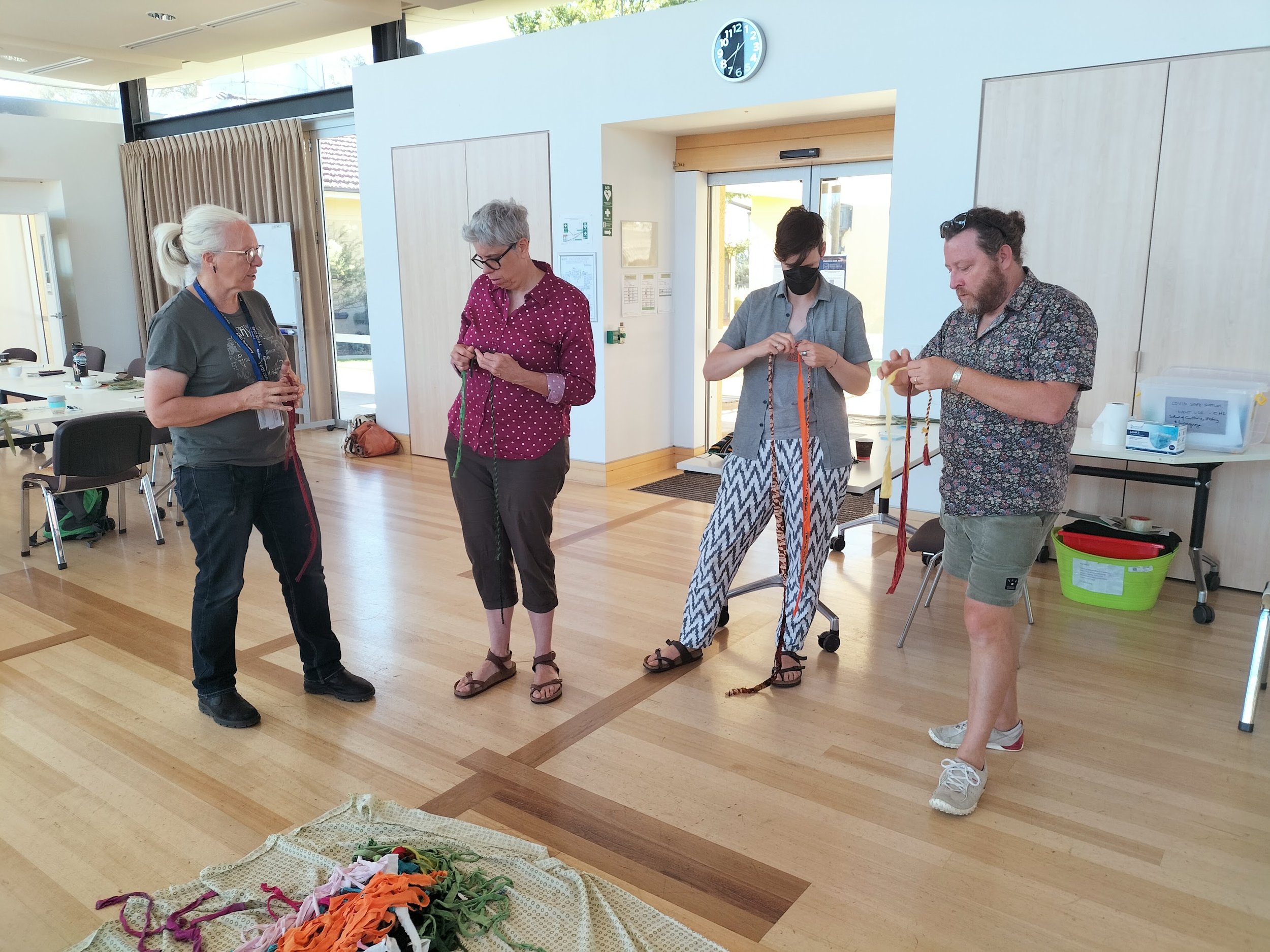 Four people standing in a room with wooden floors, looking at colorful fabric scraps on a cloth on the floor. One person has a lanyard and glasses, two are holding strips of fabric, and one is holding a striped fabric. The room has large windows, a clock showing 12:04, and a table with chairs.