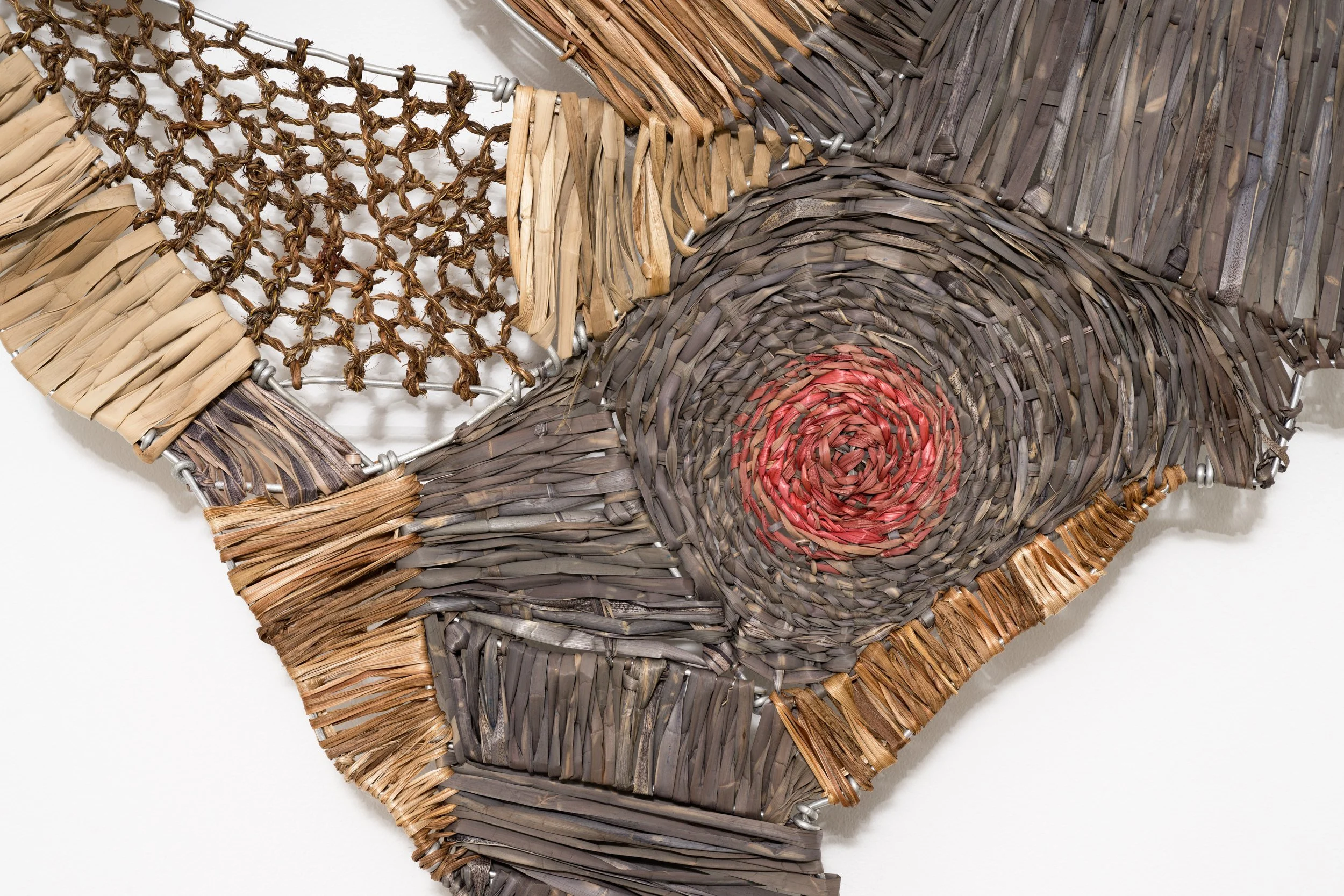Close-up of a colorful woven artwork made from various natural and dyed plant fibers, including tan, gray, and red hues, arranged in geometric and circular patterns.