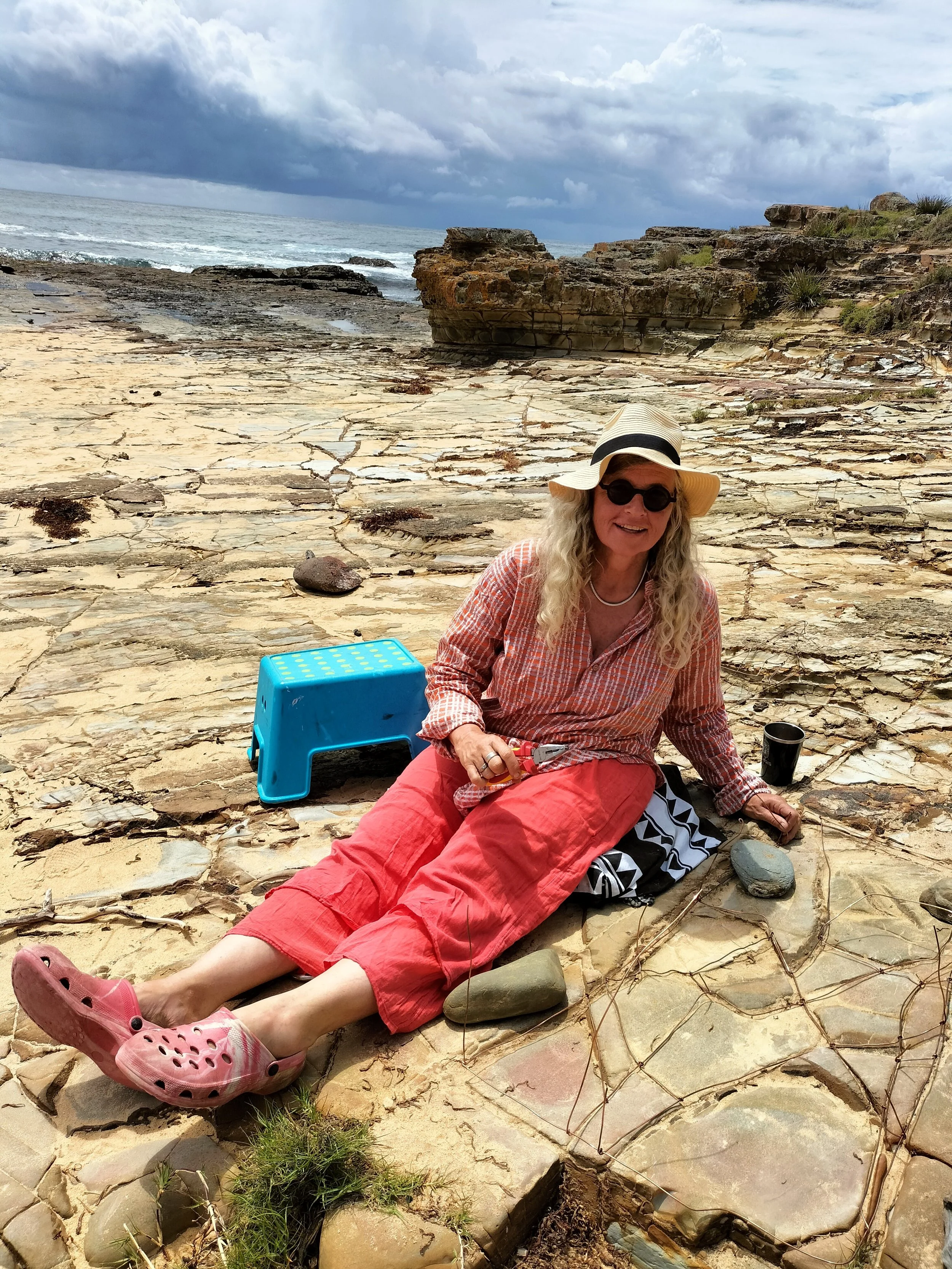 A woman with long blonde hair, wearing a straw hat and sunglasses, is sitting on a rocky beach with her legs stretched out. She is smiling and holding a tool. There is a blue plastic step stool and a black cup next to her. The beach has cracked, layered rocks, and in the background, the ocean with small waves and a cloudy sky are visible.