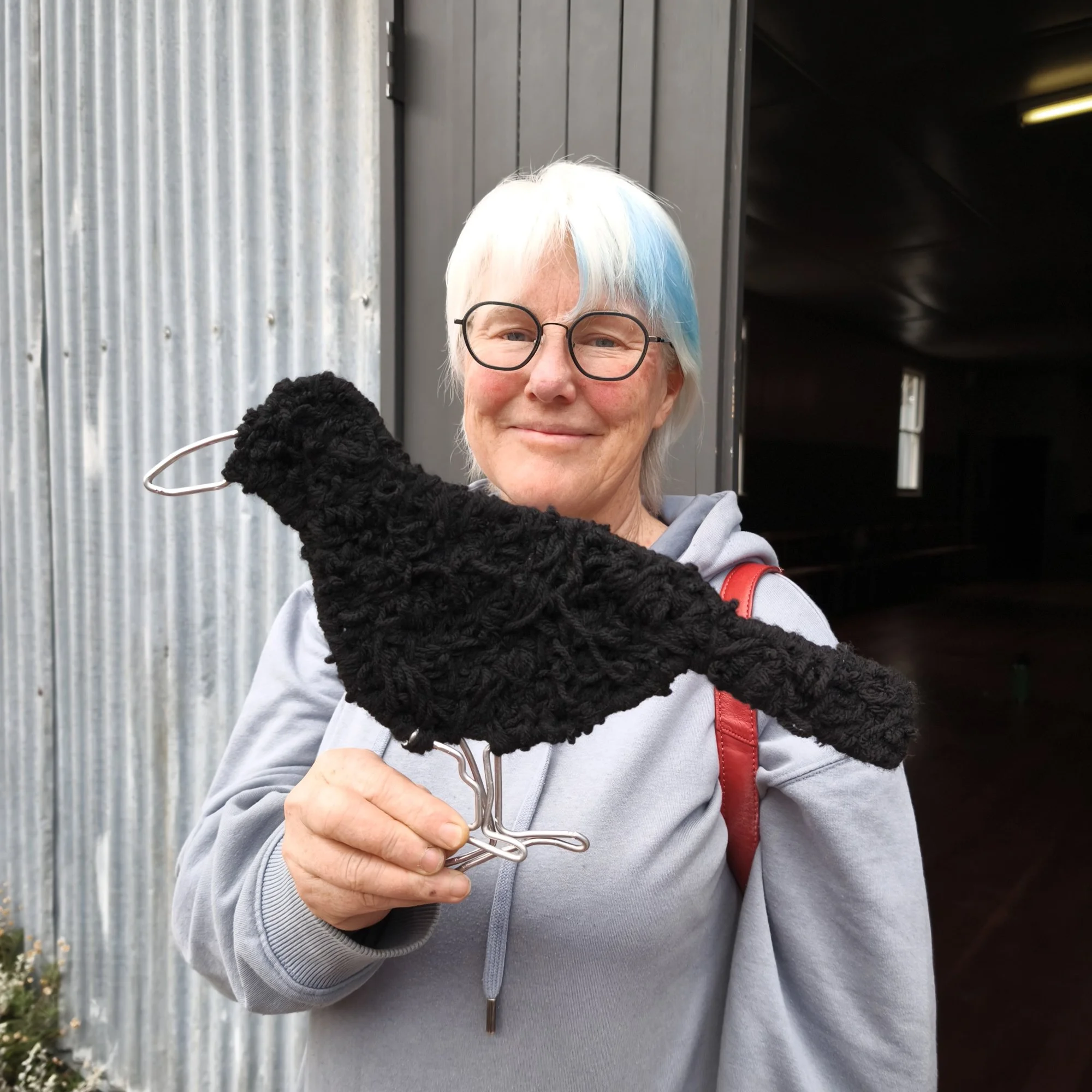 Person with grey hair and glasses holding a black fibre sculpture resembling a magpie infront of a metallic wall and a dark building.