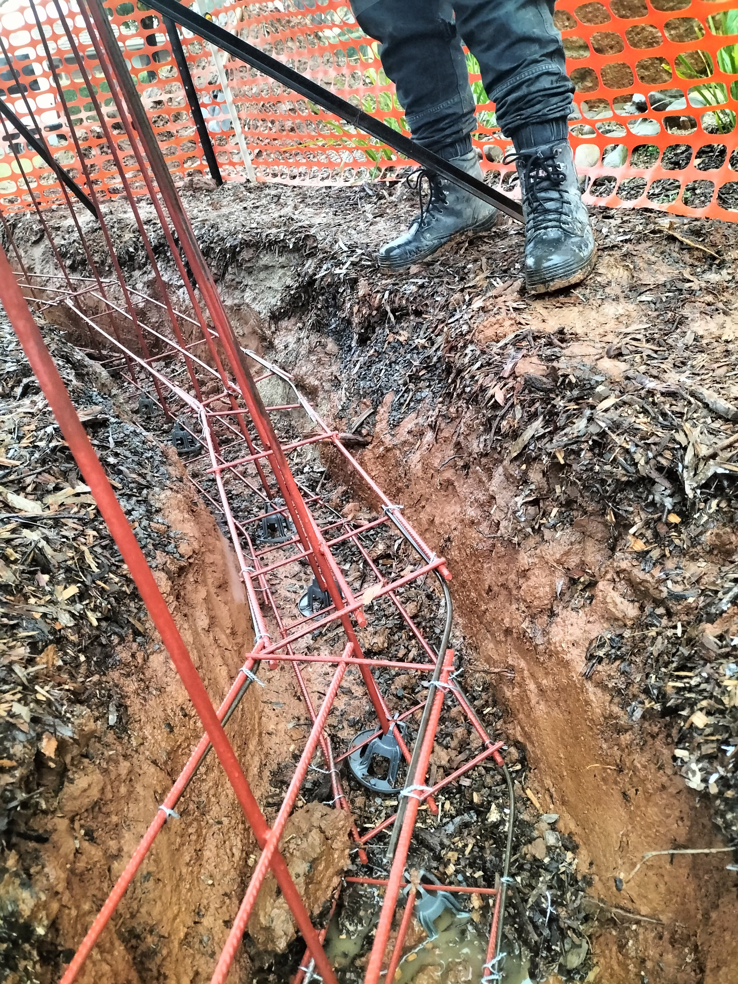 A person wearing combat boots and dark pants working on underground utility cables with a metal trench box and orange safety barrier at a construction site.