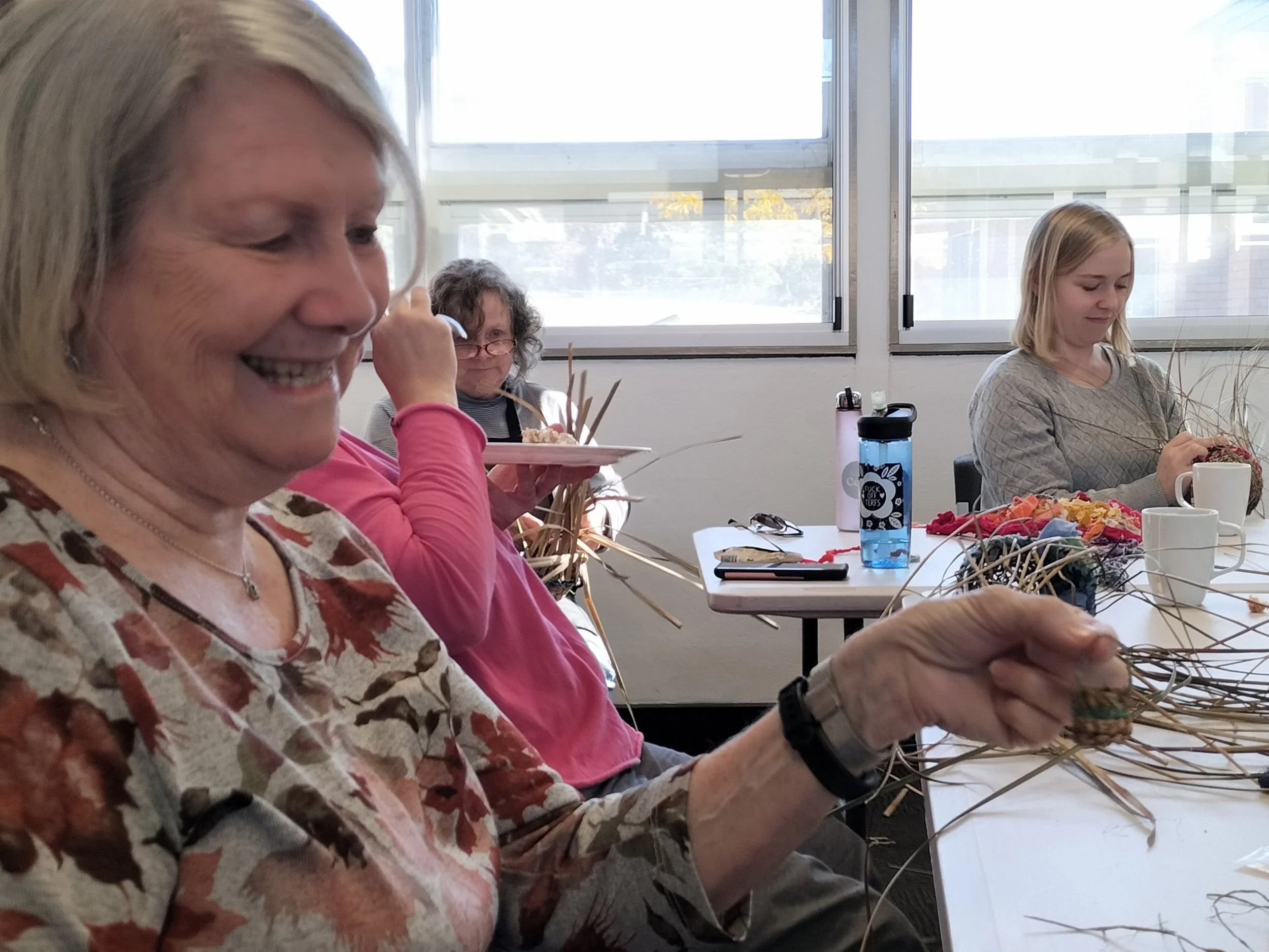A group of women sitting at a table, engaging in a craft activity involving weaving or basket-making, with various supplies on the table including a water bottle with a sticker and some yarn or string.