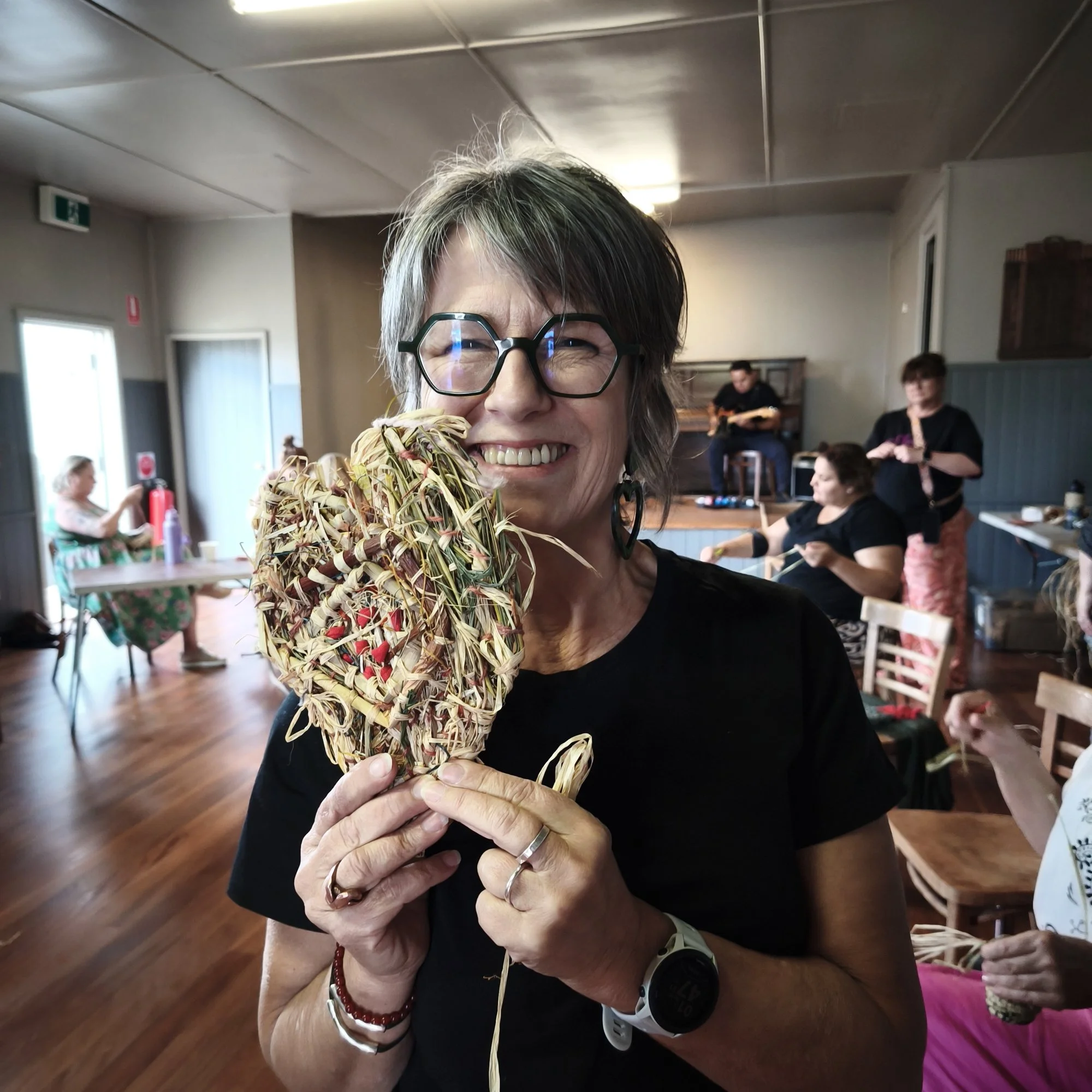 A woman with short grey hair, glasses, and wearing a black shirt is holding a large, intricate, woven heart sculpture made from raffia, smiling at the camera in a casual indoor setting with other people visible in the background.