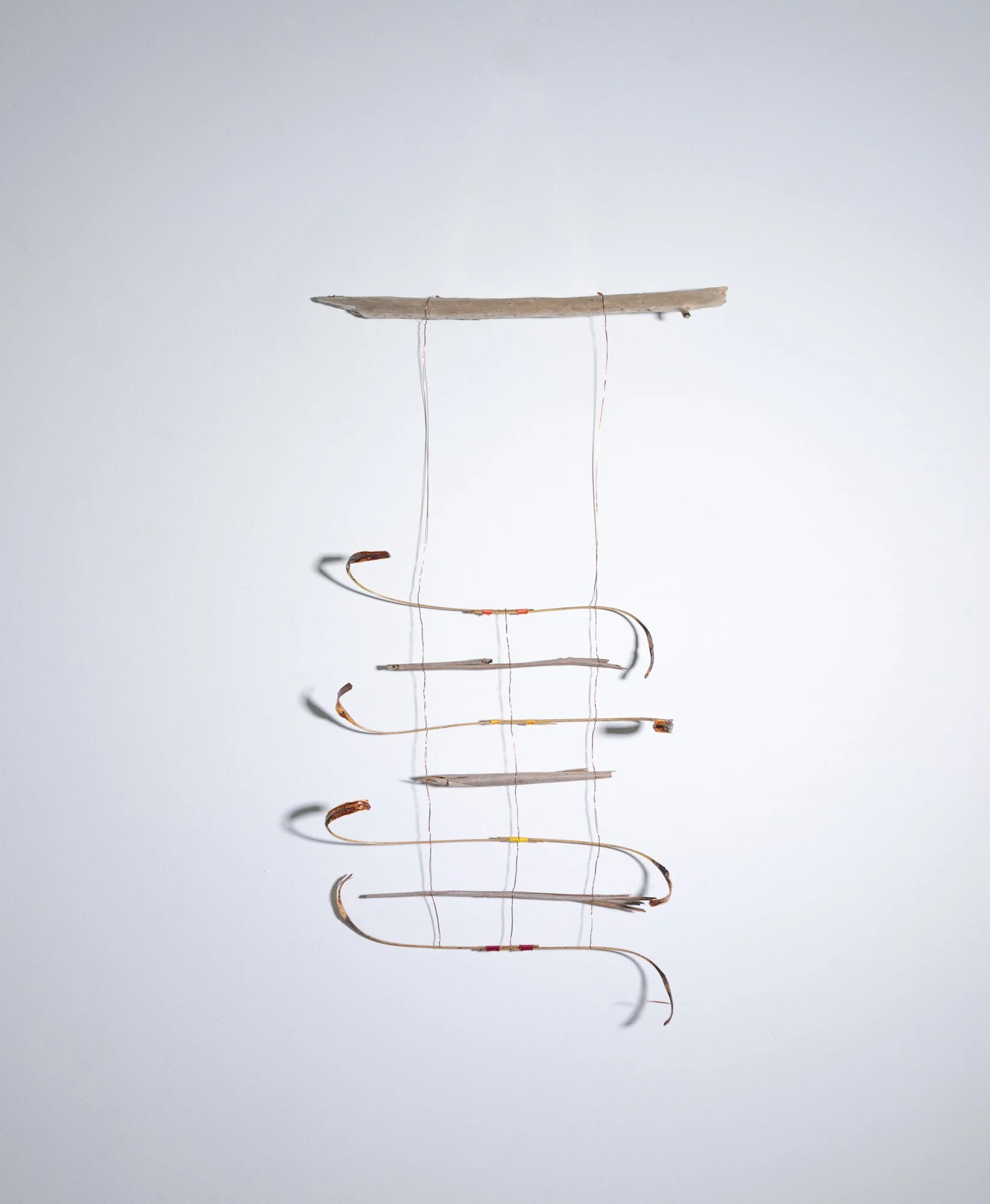 A minimalist art installation with a horizontal piece of weathered wood suspended by thin wires against a white wall, with black shadows cast below.