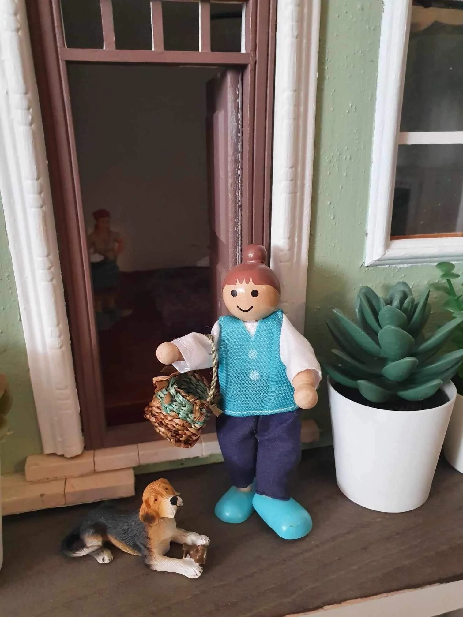A decorative doll of a girl standing in front of a door from a doll's house with several small dogs nearby, a green wall, a window, and a large plant in a white pot. She holds a miniature twined basket.