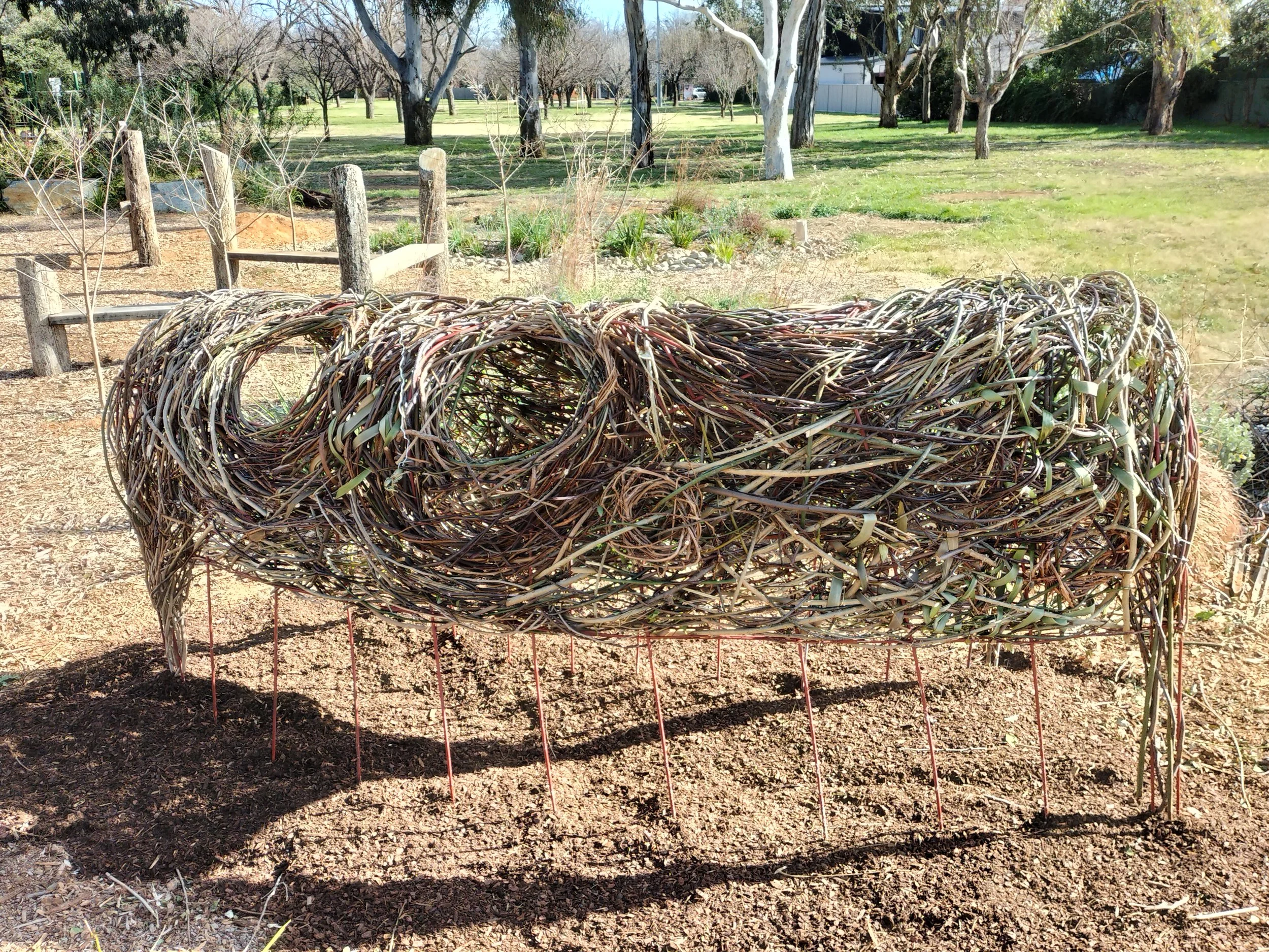 A woven tunnel-like structure entitled 'The Bowery' made of twigs and branches is placed in a park with green grass, trees, and a clear blue sky in the background.