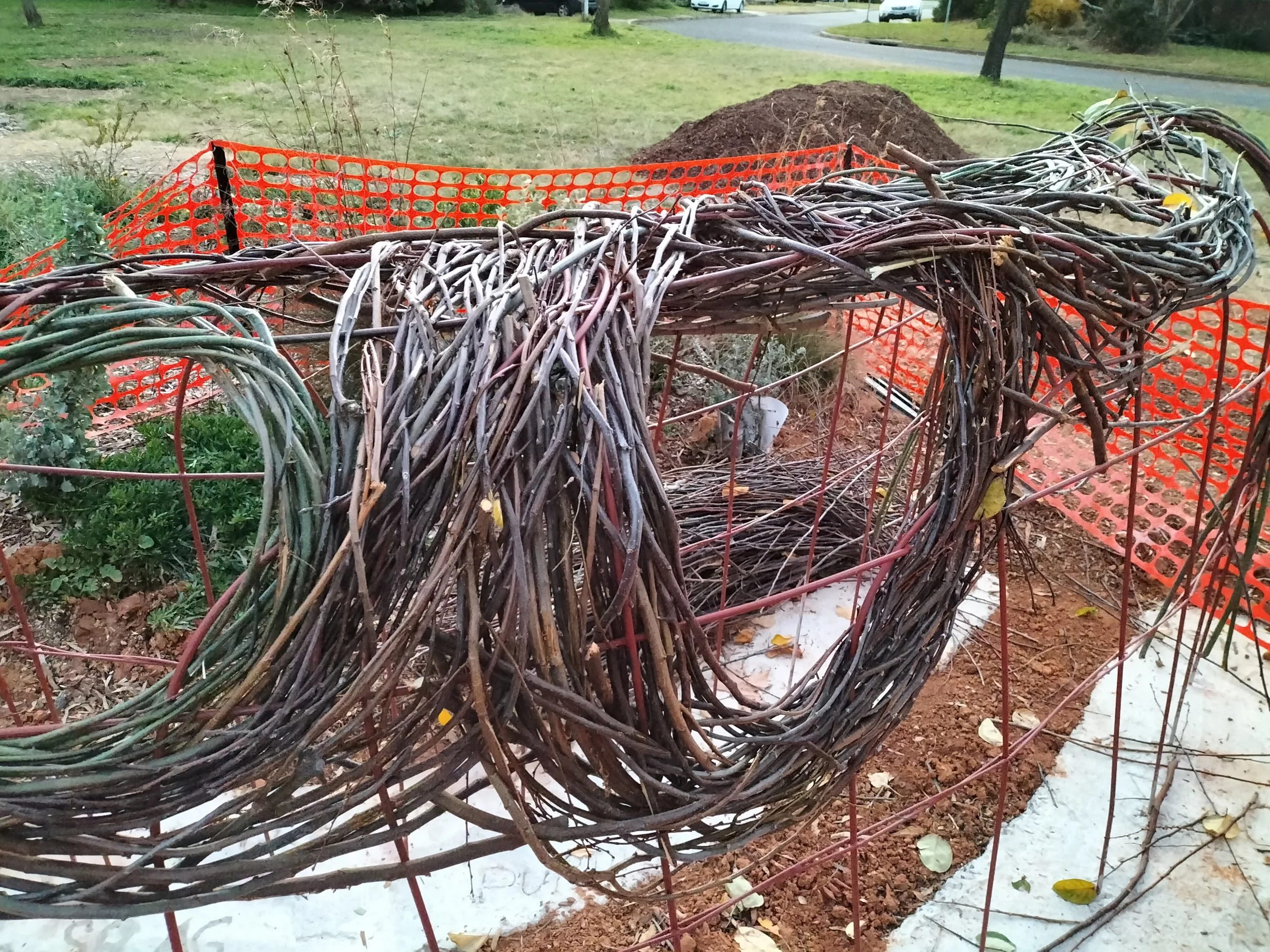 A wooden structure made of intertwined branches, surrounded by bright orange construction fencing, in a grassy outdoor area with a pile of dirt and a sidewalk in the background.