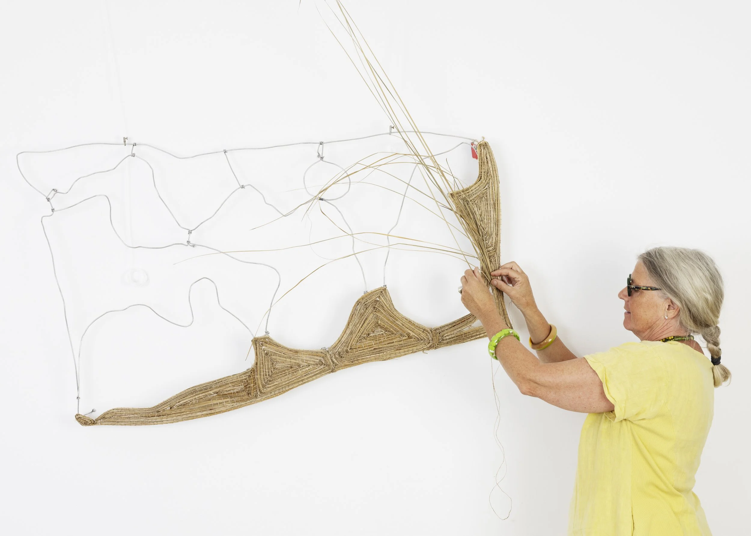 An older woman with gray hair, wearing glasses and a yellow top, is attaching a woven or wicker star-shaped decoration to a wall that features a wire star and constellation-like display.