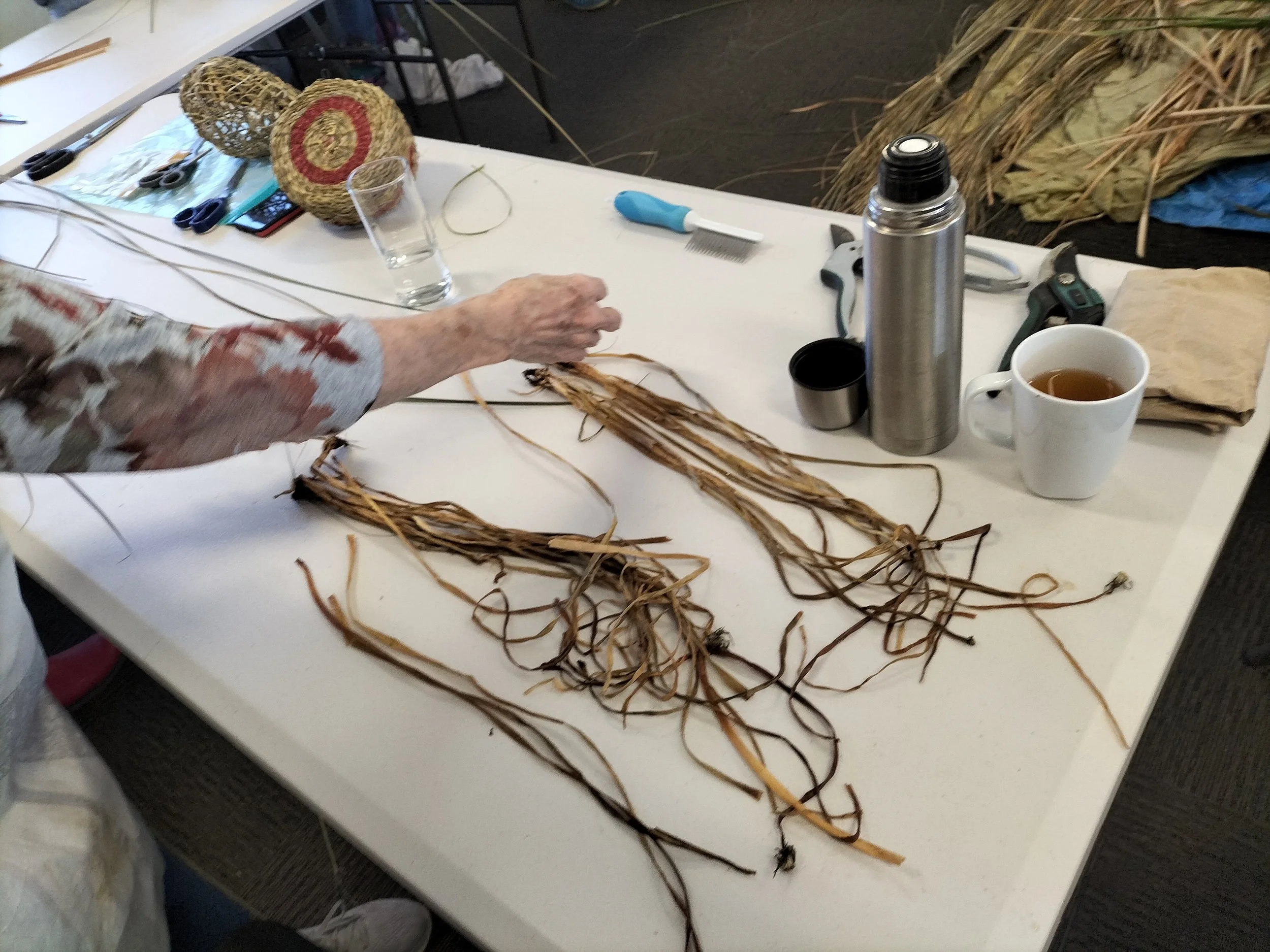 A person working with dried plant fibers on a white table, with crafting tools, a glass of water, a thermos, and a mug of tea nearby.