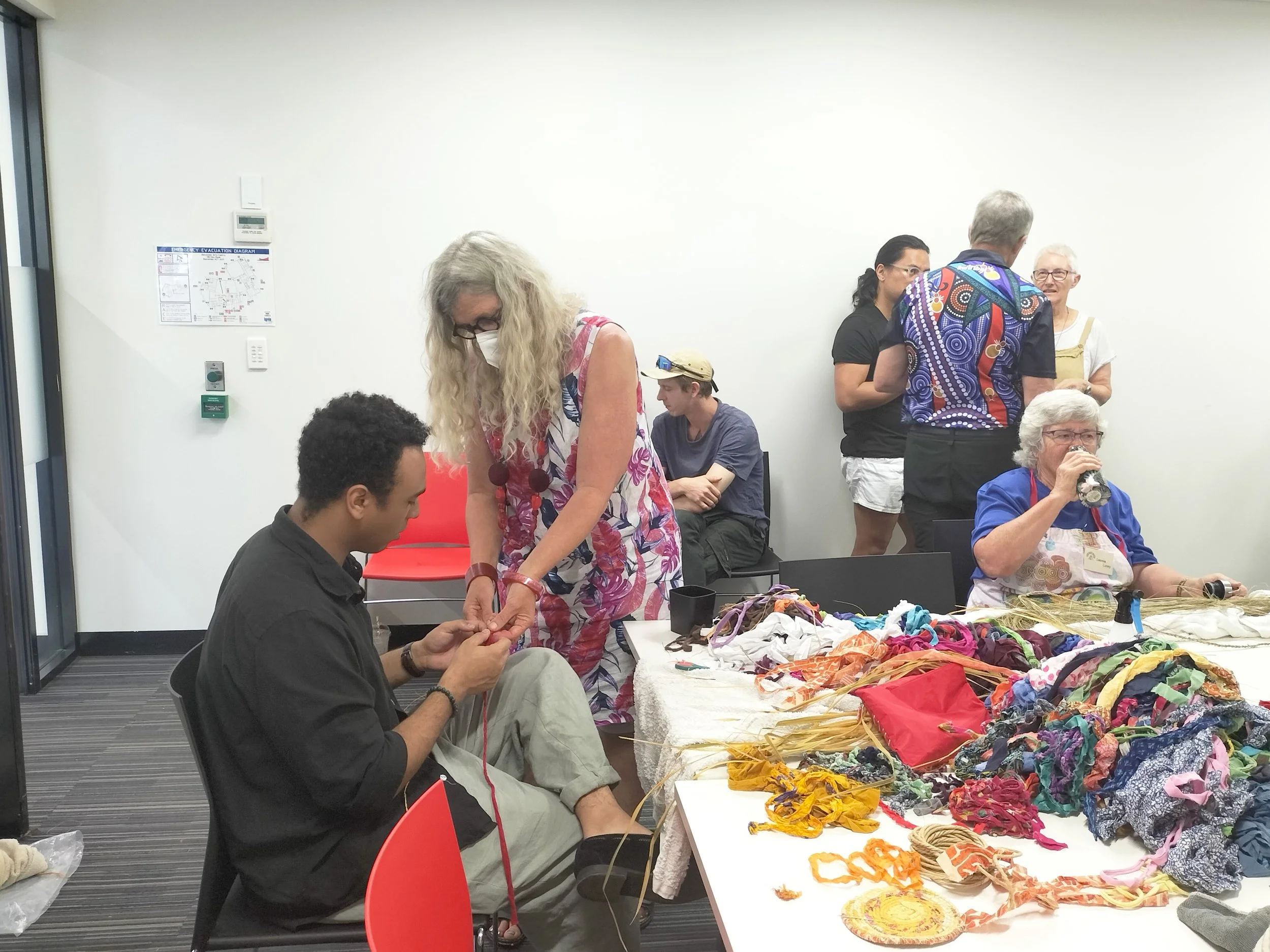 People engaged in crafting activities at tables with colorful fabric and craft supplies in a room with plain white walls.