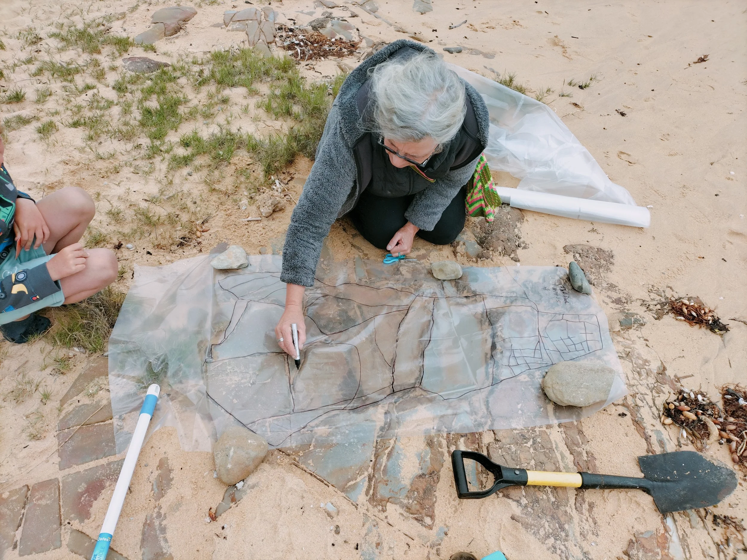 An older woman with gray hair wearing glasses, kneeling on sandy ground and working on a large piece of paper with black marker lines, surrounded by rocks, plastic, and tools.