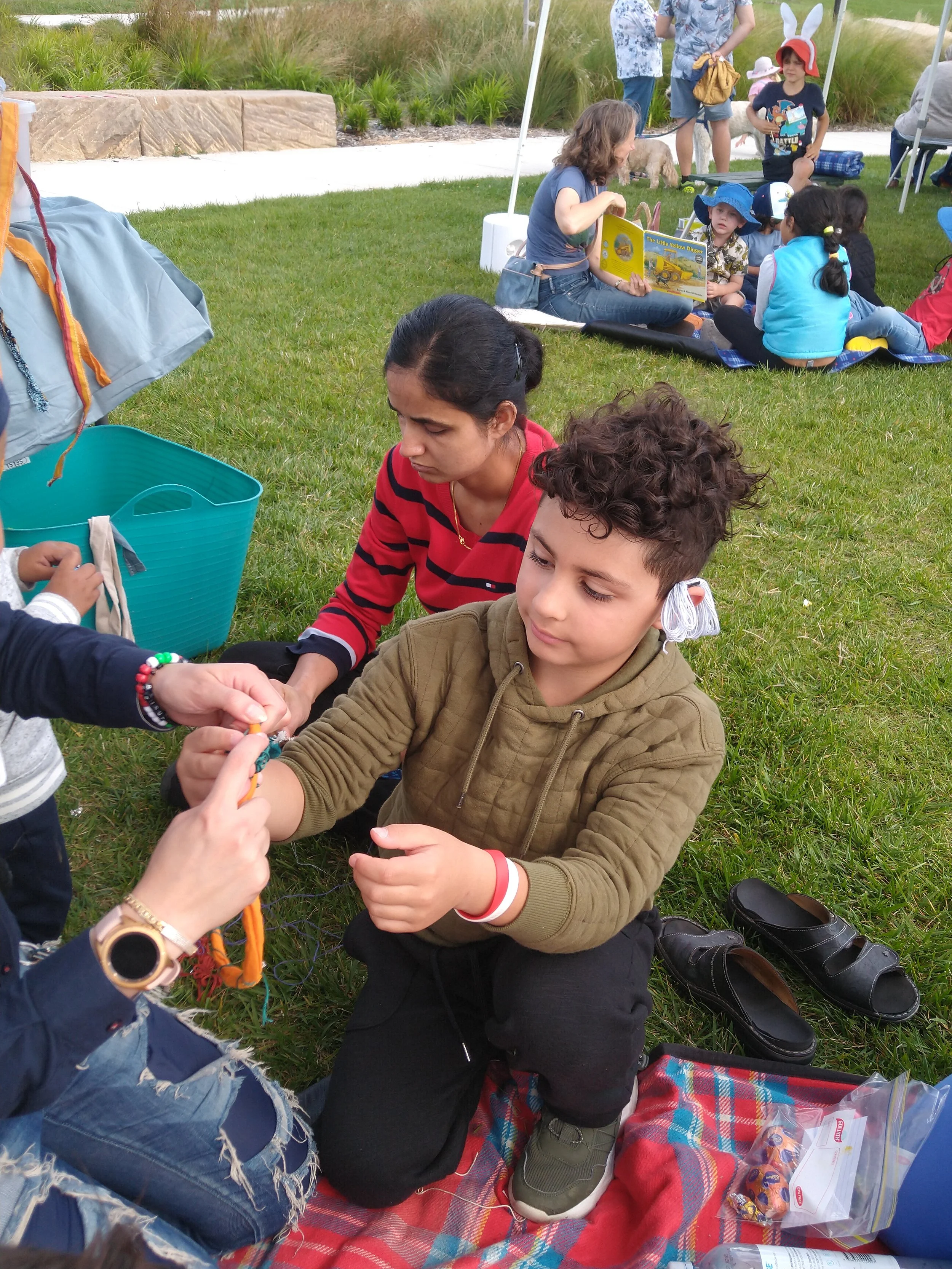 Children and adults sitting on a red and blue plaid blanket outdoors, with children engaging in an activity and adults watching, some children wearing costumes or hats, and a book titled 'The Little Yellow Digger' visible in the background.