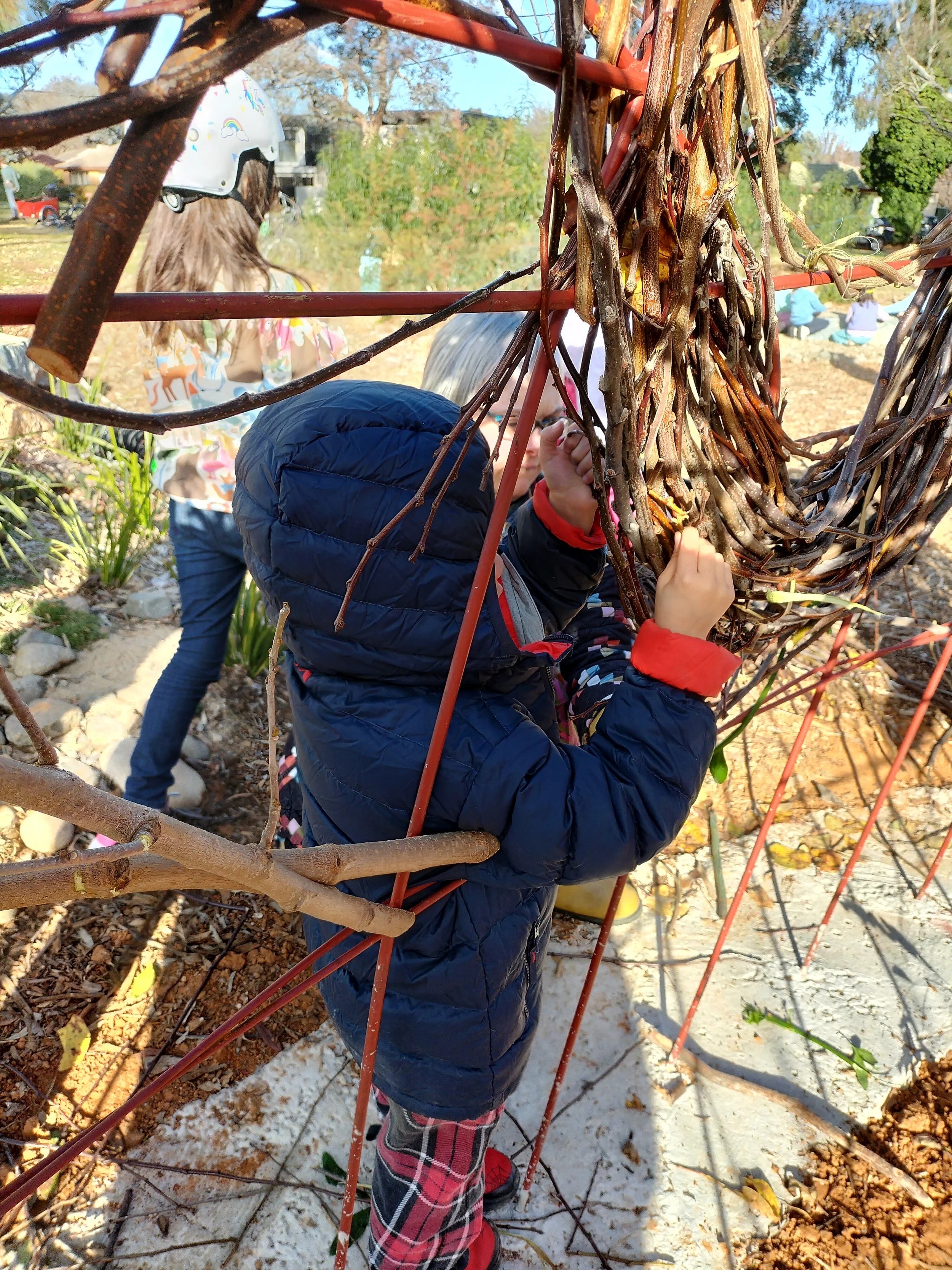 Children playing outdoors, climbing and exploring among brown tree branches with some snow on the ground, wearing jackets, pants, and boots on a sunny day.