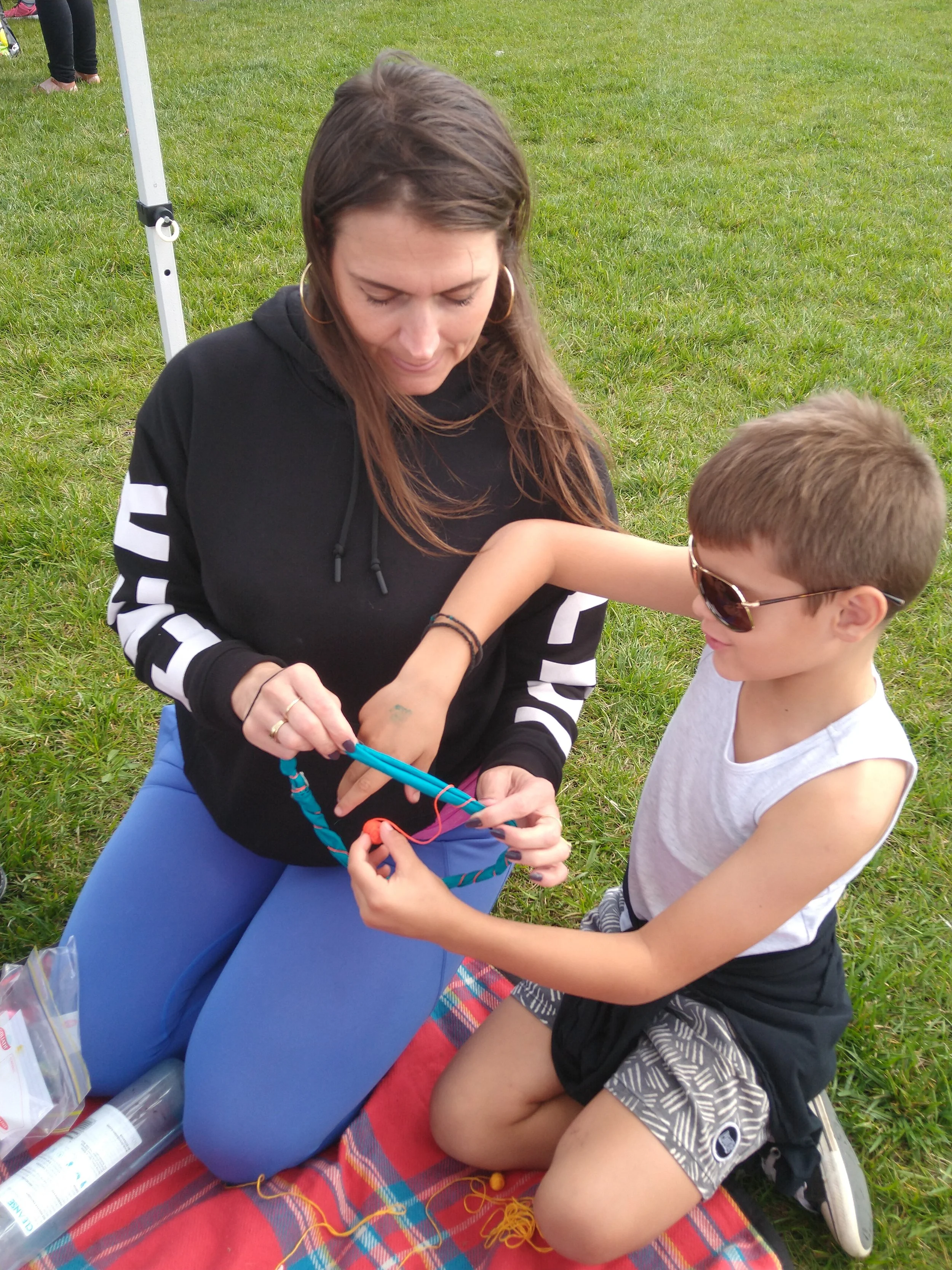 A woman and a boy sitting on a blanket outdoors. The woman is holding tangled yarn and a crochet hook, while the boy is watching. They are sitting on a red plaid blanket on the grass.