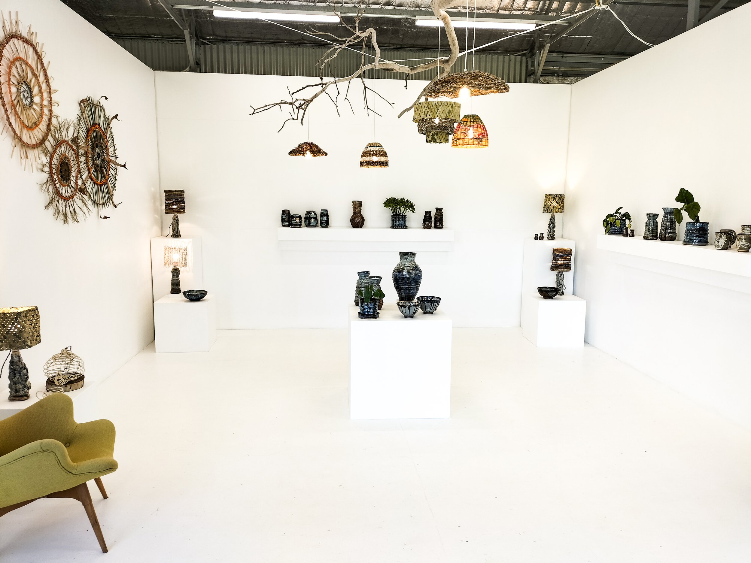 An art display in a white-walled gallery featuring woven basket wall hangings, a branch decor suspended from the ceiling, and various ceramic vases and sculptures arranged on white pedestals and walls, with two lamps providing soft lighting.