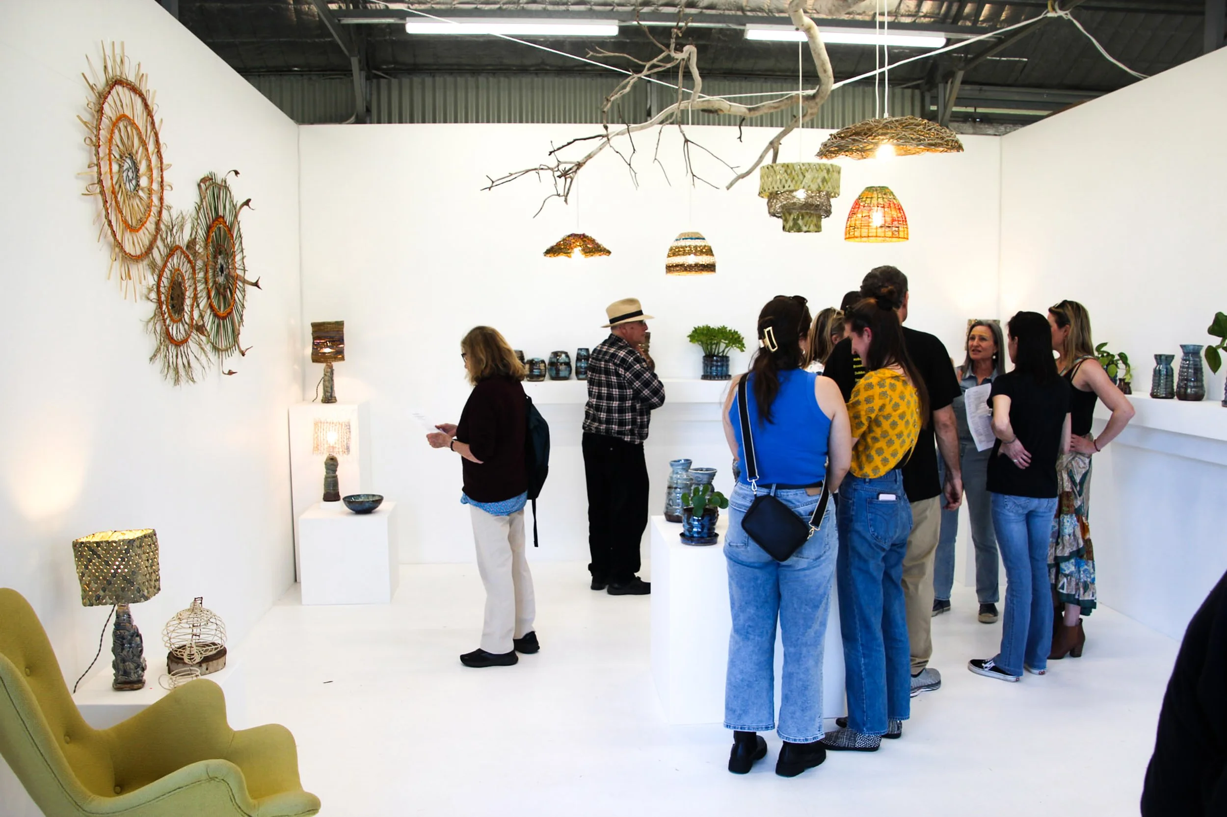 People gathered in an art gallery or craft exhibition, viewing and discussing various handcrafted items, including colorful woven and ceramic pieces, with hanging and seated décor on white walls and a branch suspended from the ceiling.