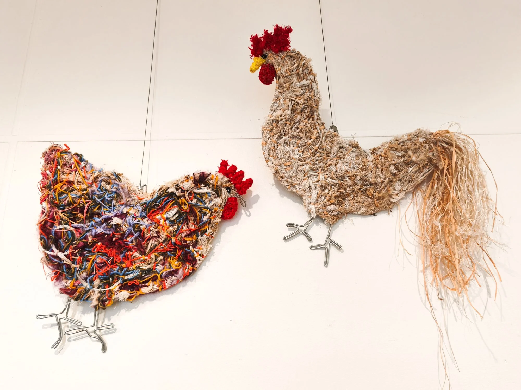 Two art sculptures resembling chickens made from intertwined and multicolored materials hung on a white wall.