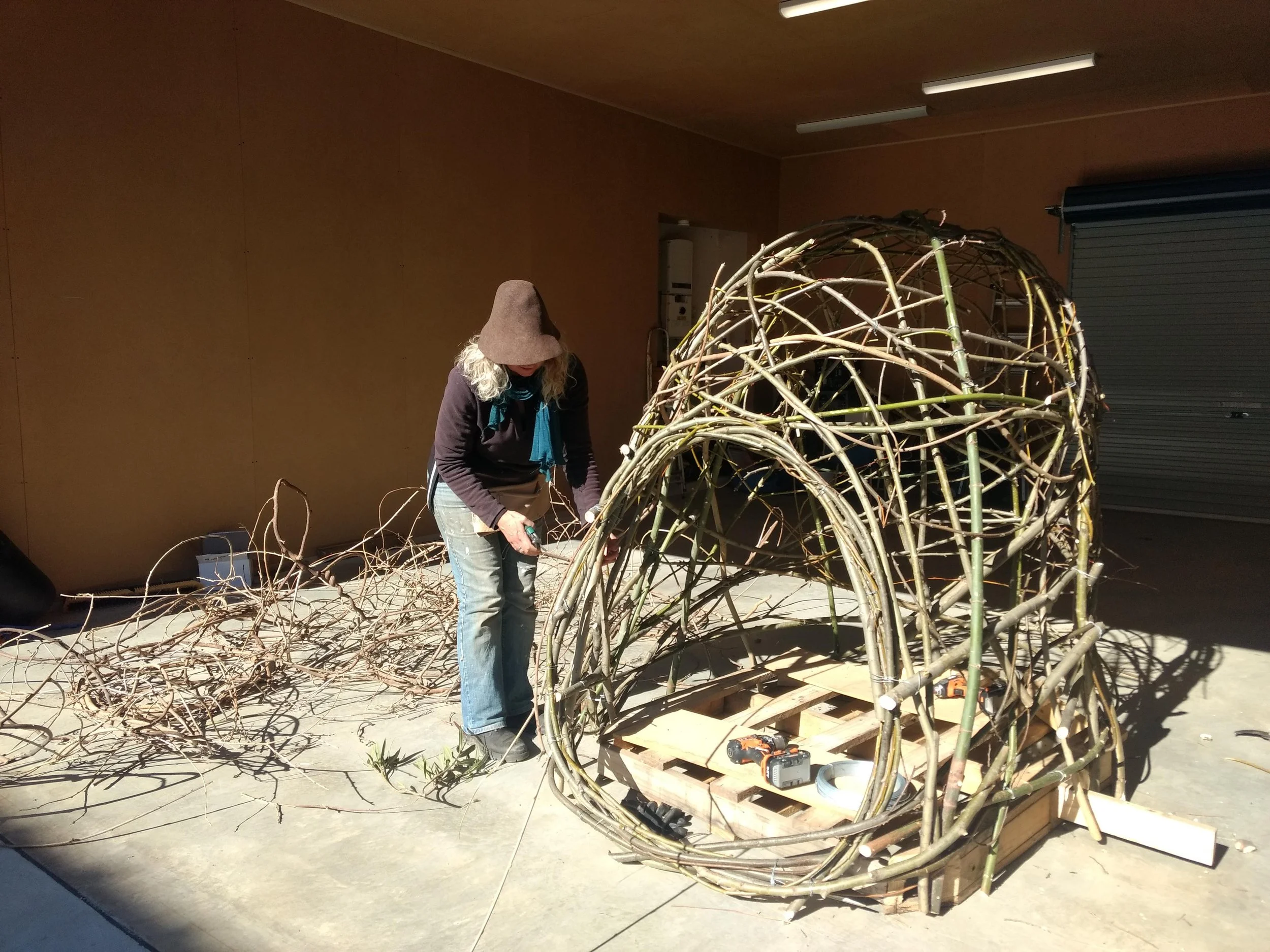 A person wearing a brown hat and blue scarf working on a large fibre sculpture made of intertwined tree branches, placed on a wooden pallet indoors.