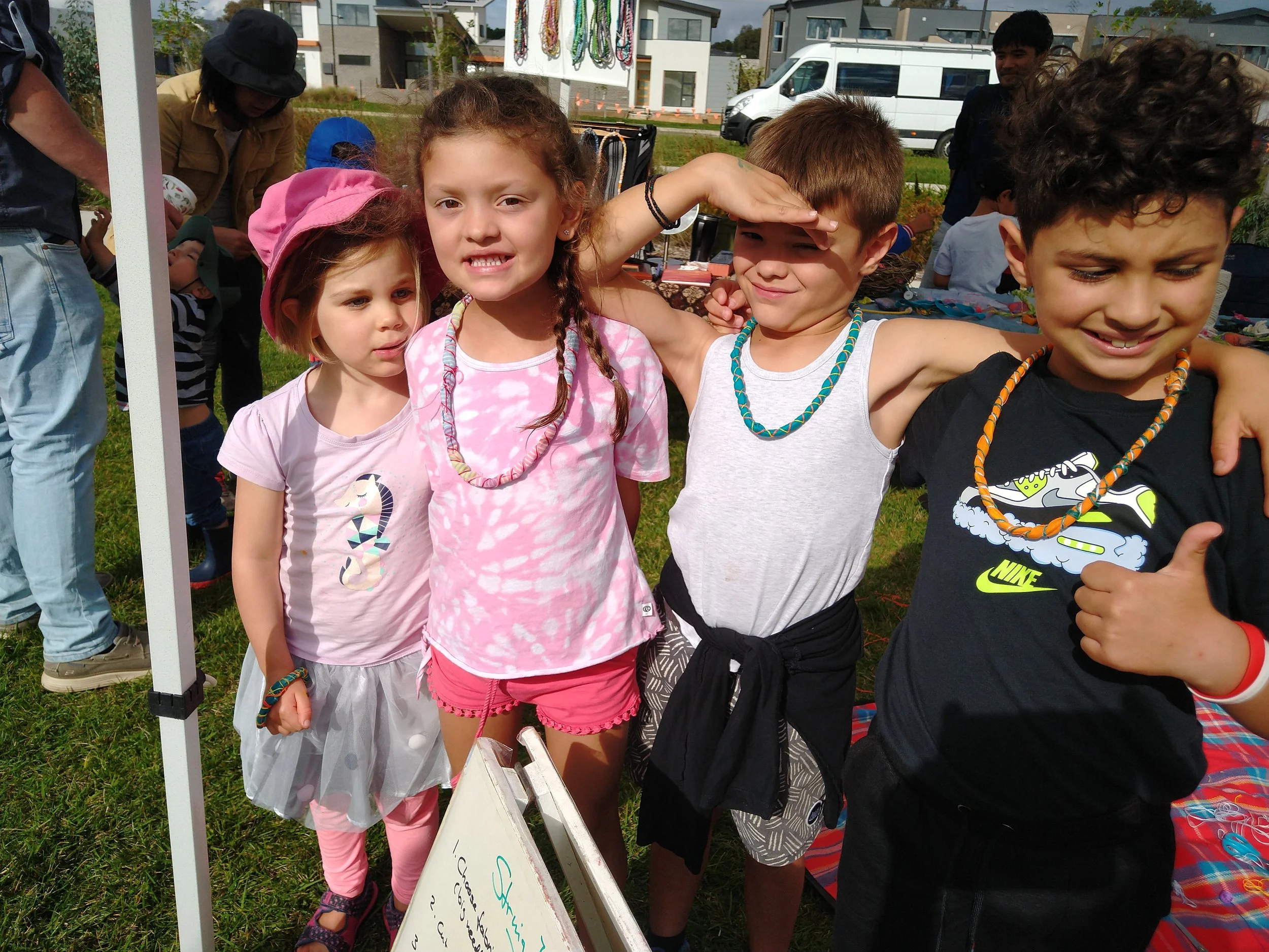 Four children standing together outdoors, smiling and wearing colorful clothing and beaded necklaces, at an event with booths and other people in the background.