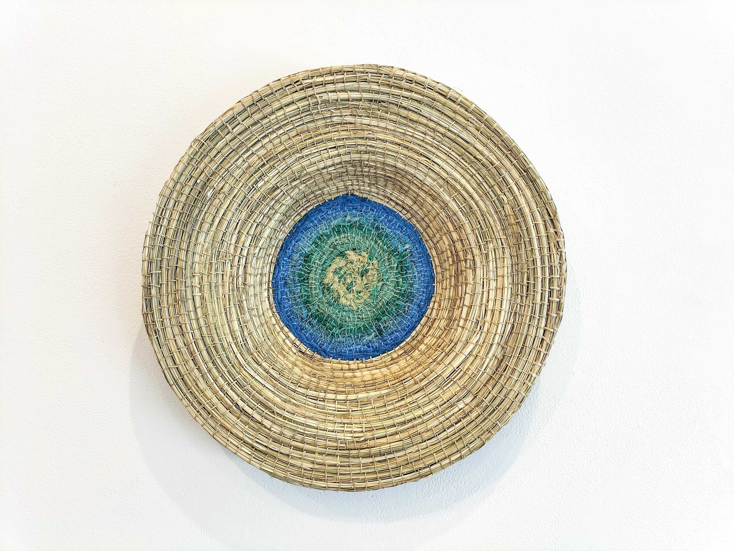 A woven decorative plate with concentric circles, featuring shades of blue, green, and beige, hanging on a plain white wall.