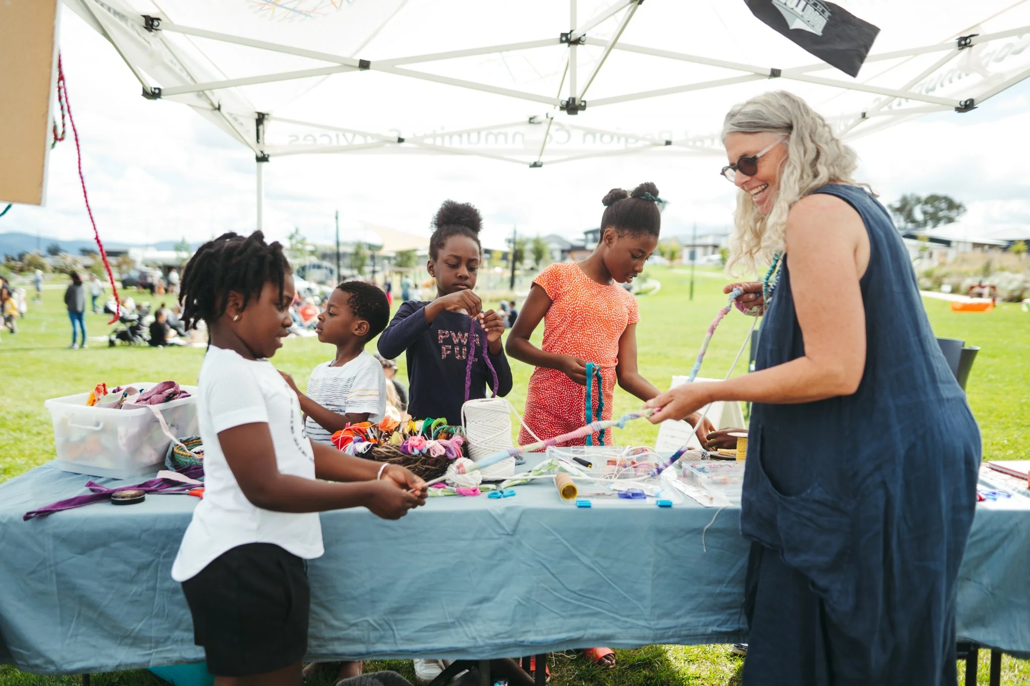 A woman with gray hair and sunglasses is selling handmade jewelry at an outdoor market stall under a white canopy. Several children are browsing the jewelry and craft items on the table. In the background, people are gathered on a grassy area, with some sitting and others walking.