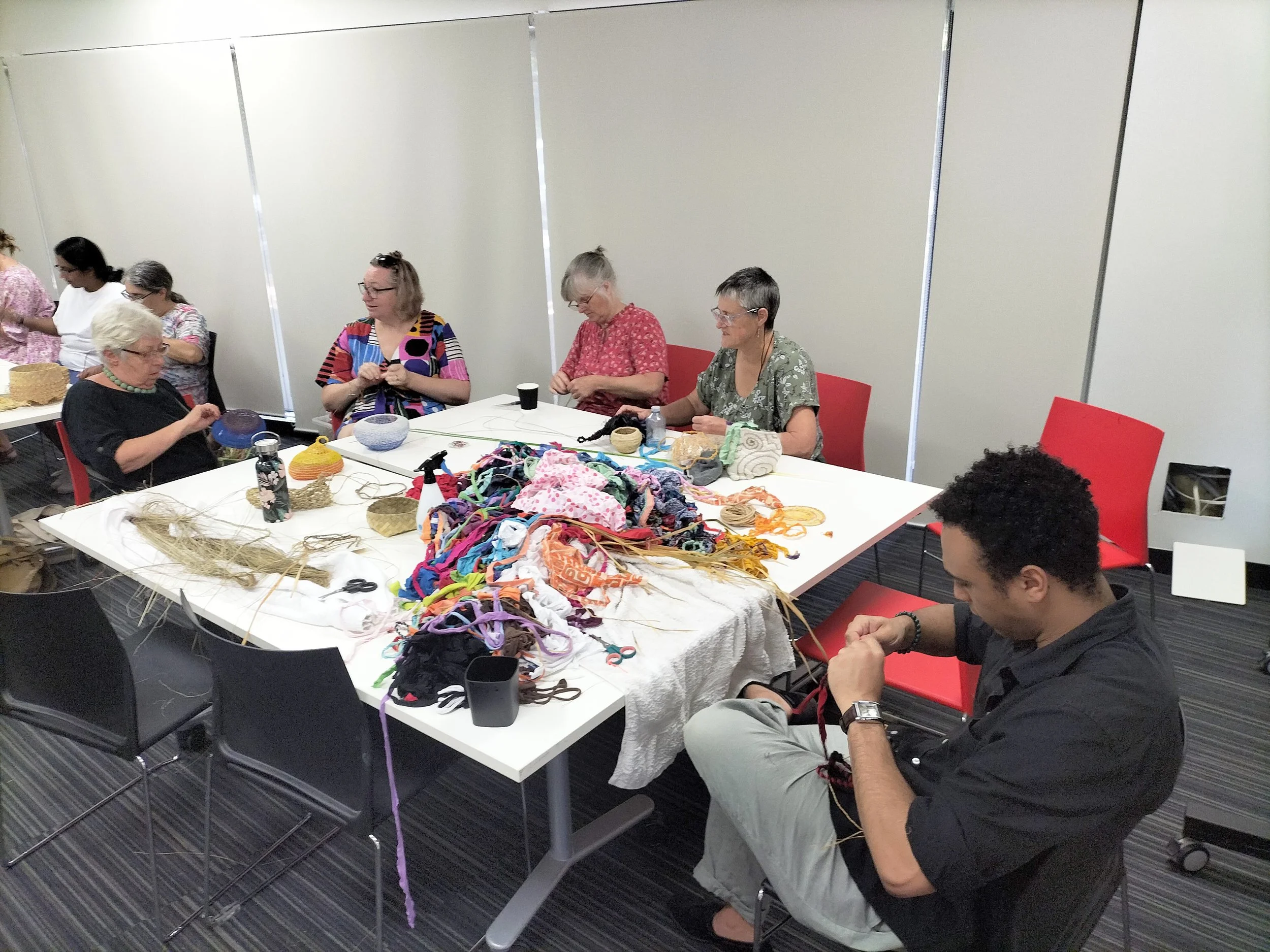 Group of people sitting at tables working on crafts or knitting, with colorful yarn and materials scattered on the tables. The room has plain white walls and gray carpet, and the people appear engaged in their activity.