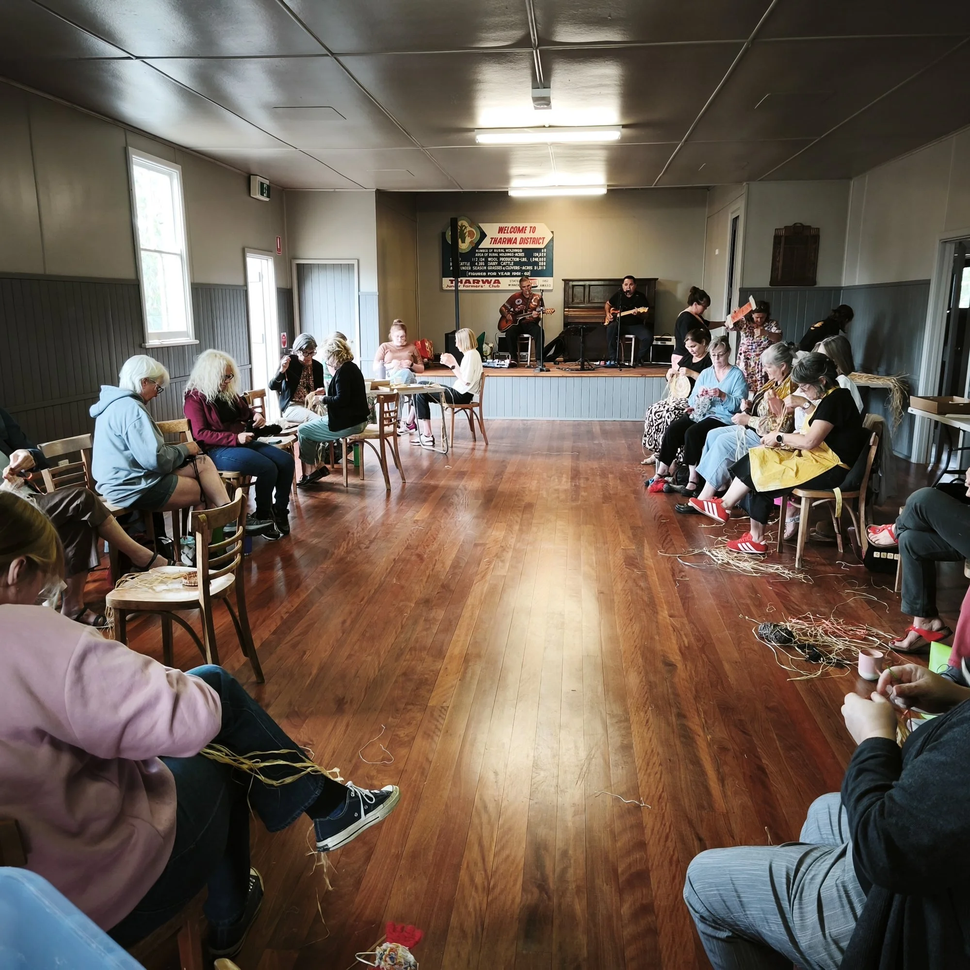 A community gathering with people sitting on chairs and the wooden floor, some using sewing needles. A stage with musicians playing guitars is at the front. There is a welcome sign for Tharwa District on the wall behind the stage.