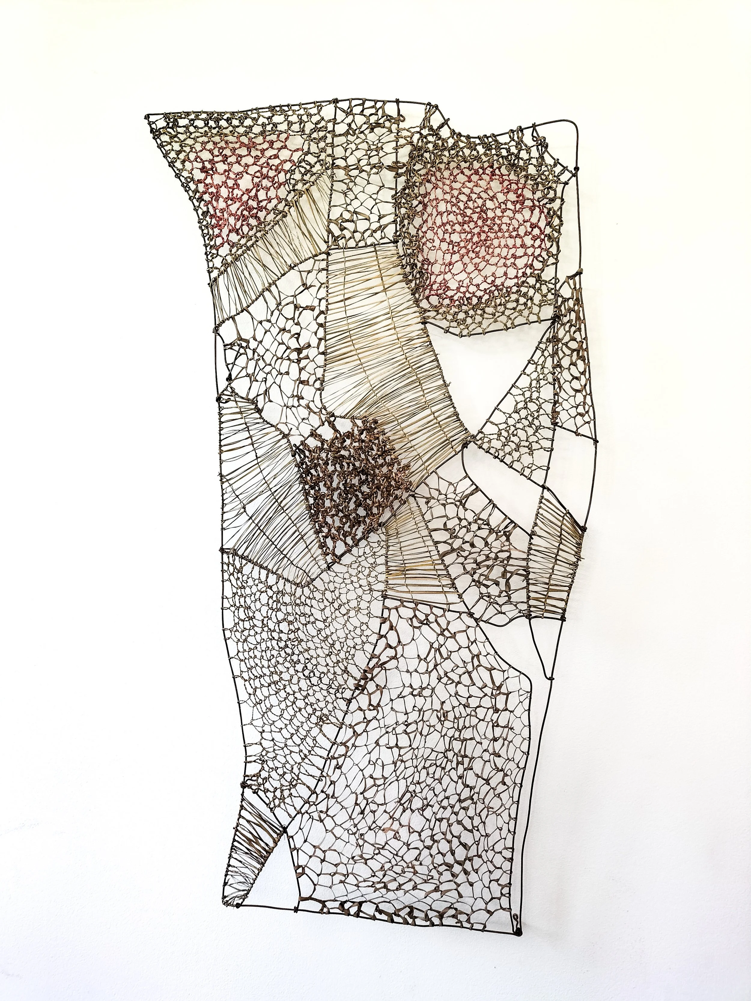 Wire sculpture resembling a stylized human figure with intricate web-like patterns in various sections, mounted on a plain white wall.
