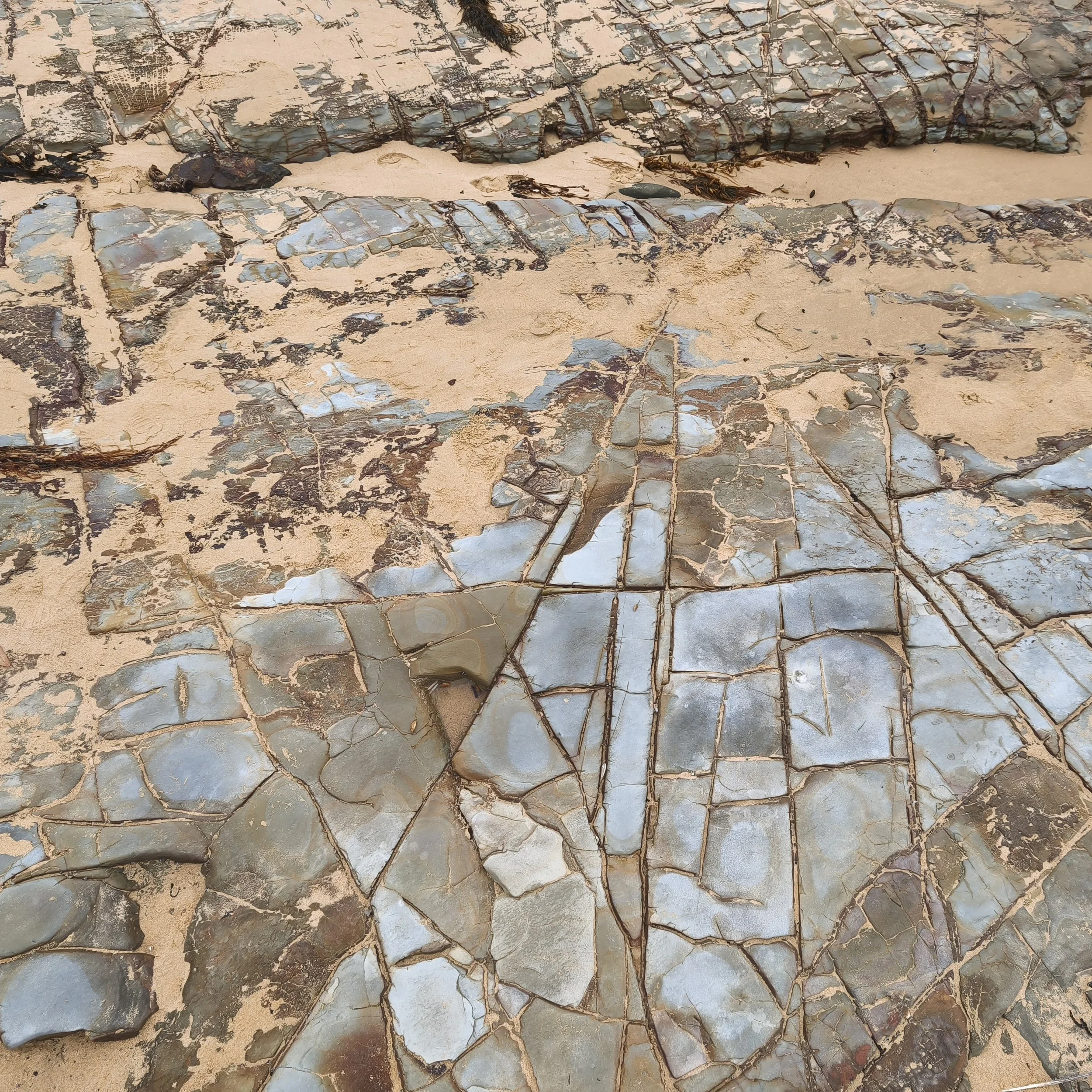 Close-up of a rock formation on sandy ground, with layers of grey and greenish stones separated by thin lines of dark veins.