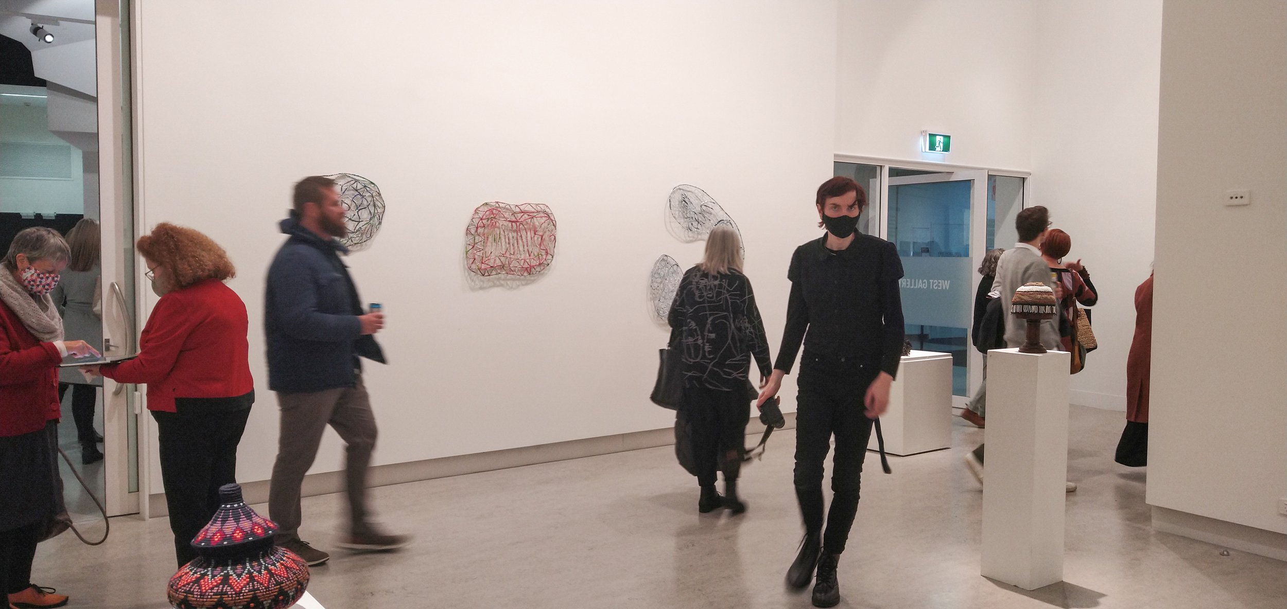 Gallery visitors observing abstract wire sculptures on white walls, some wearing masks, in a modern art museum.