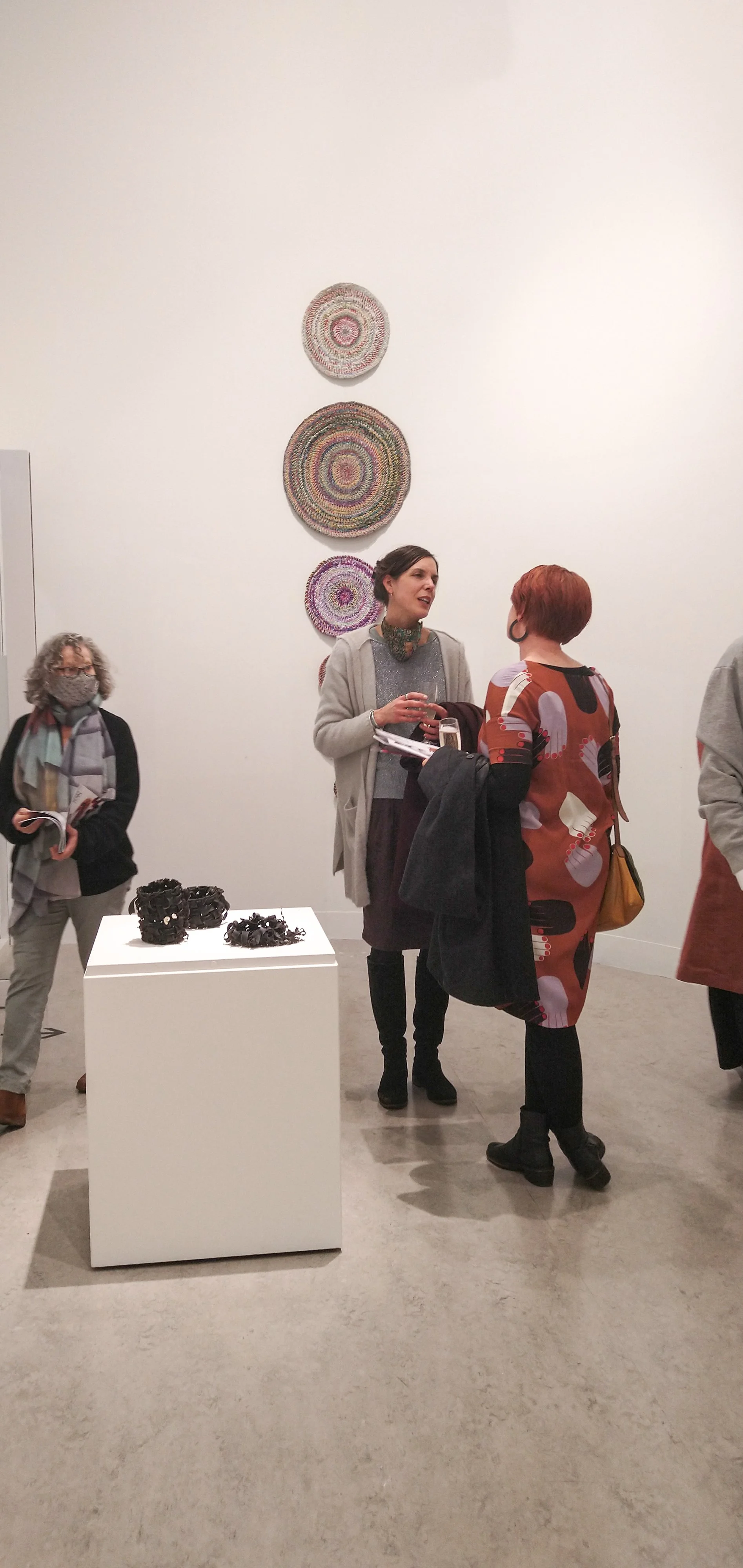People engaging in conversation at an art gallery, with colorful woven circular artwork on the white wall behind them and black sculptural pieces on a white pedestal in the foreground.