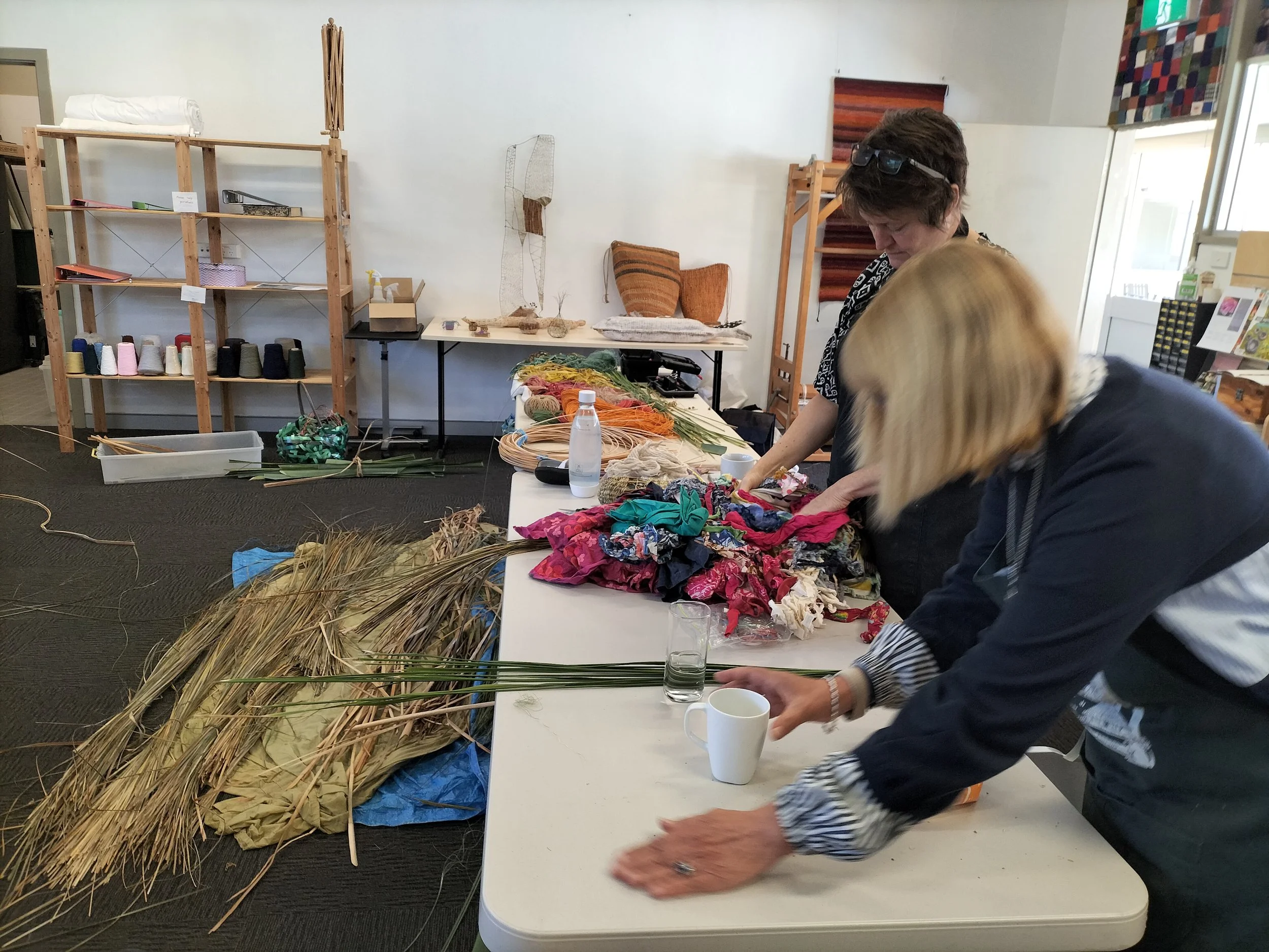 Two women working with natural materials in a room with shelves and artwork, possibly for a craft as part of a weaving workshop.