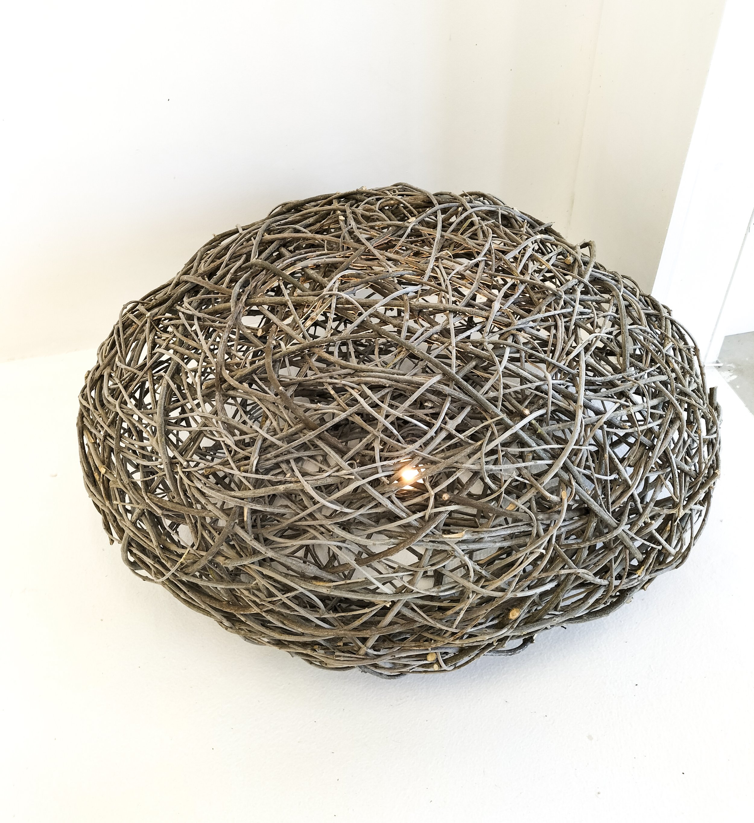 Close-up of a large, woven, spherical decoration made from intertwined twigs or vines, resting on a white surface with a plain white background.