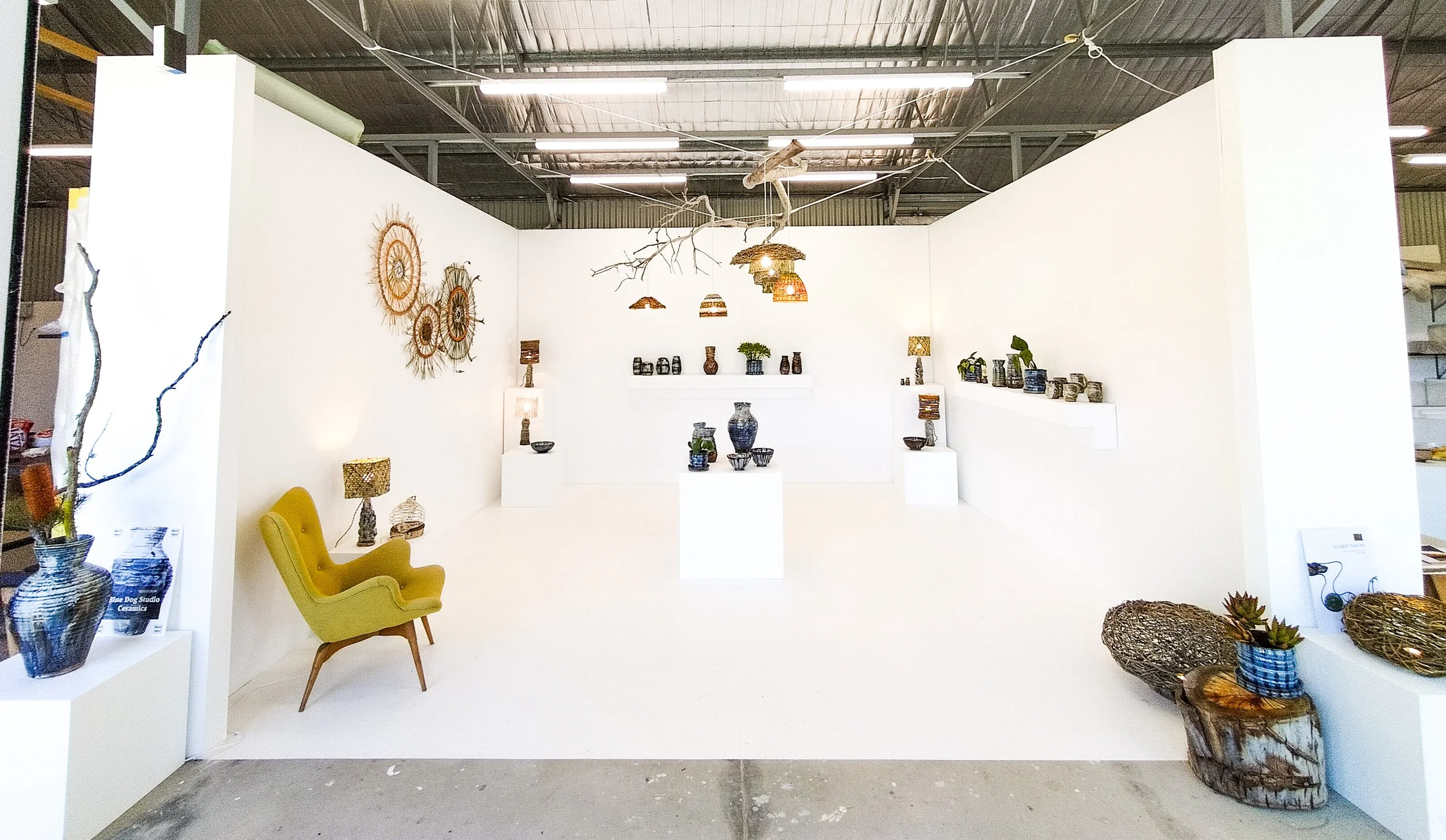An art gallery display featuring various ceramic vases, pots, and decorative items on white pedestals and shelves, with a yellow armchair and wicker baskets in the foreground and wall art on the walls.