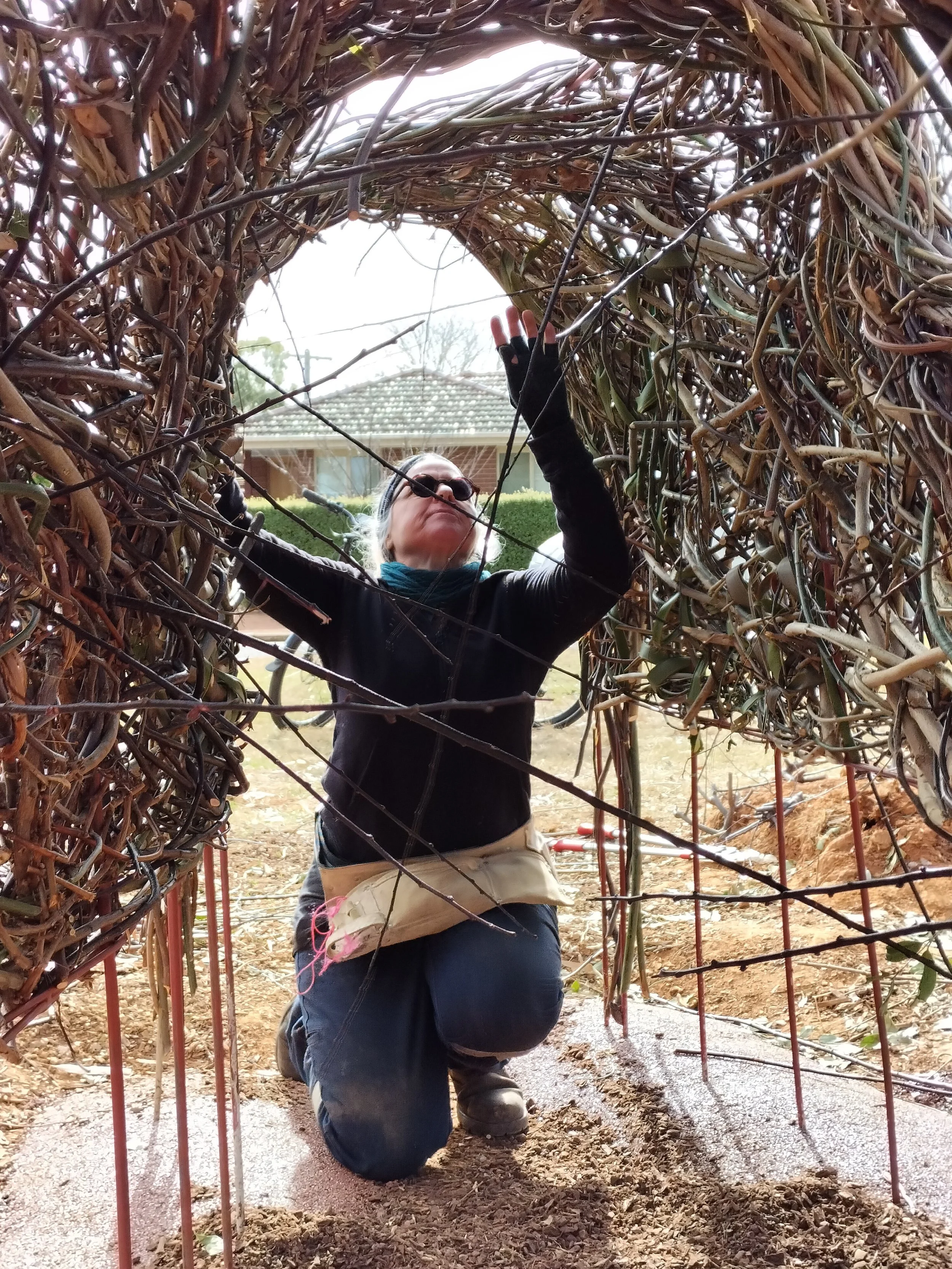 Person kneeling in a garden bed, working under a large woven arch made of intertwined branches and twigs. The person is wearing glasses, a black jacket, and beige pants, and is reaching up with one hand pulling sticks through while looking upwards.
