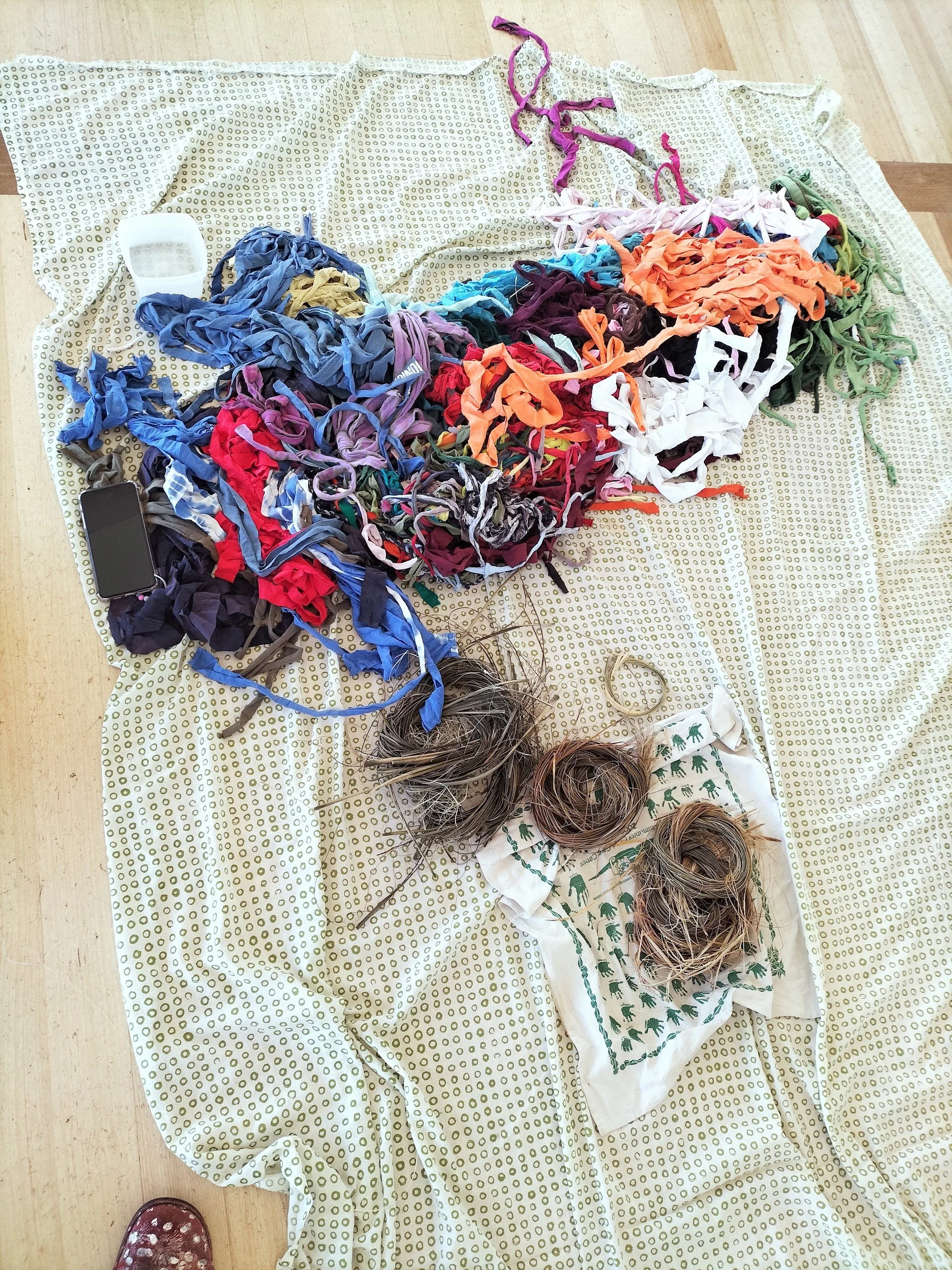 Colorful tangled strips of fabric or t-shirts on a beige sheet with a polka dot pattern, some hair coils, a white bag with green pattern, and other items on a light wood floor.