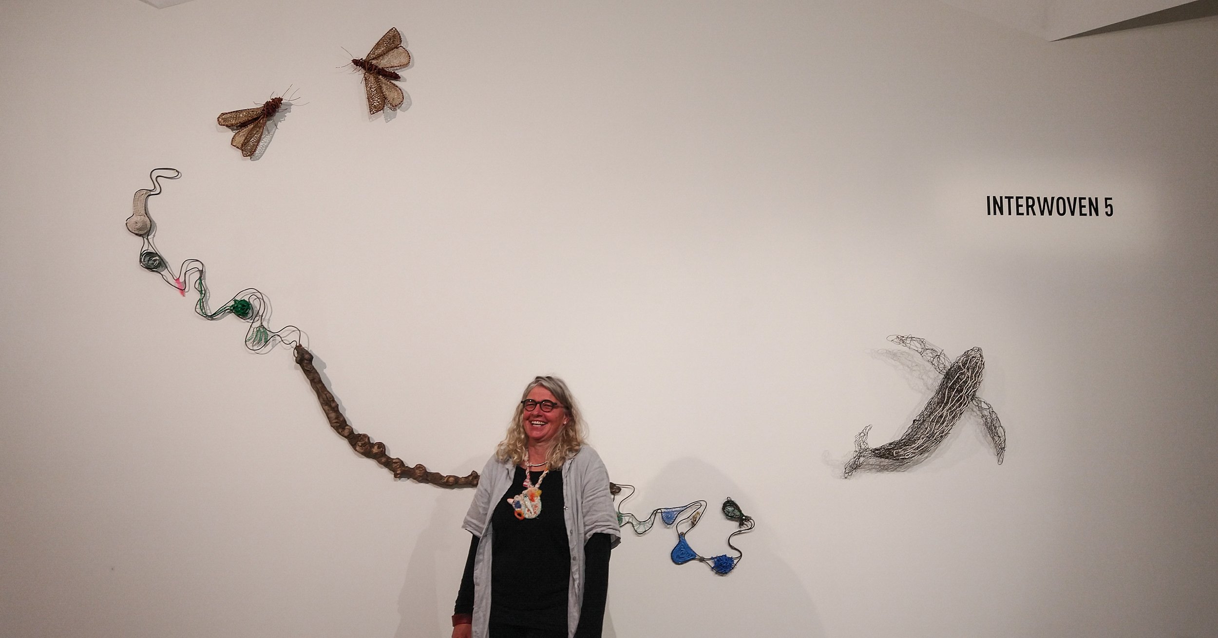 A woman with glasses and long blonde hair laughing in front of a white wall with abstract art made of intertwined wires and objects, including butterflies and labeled 'Interwoven 5'.