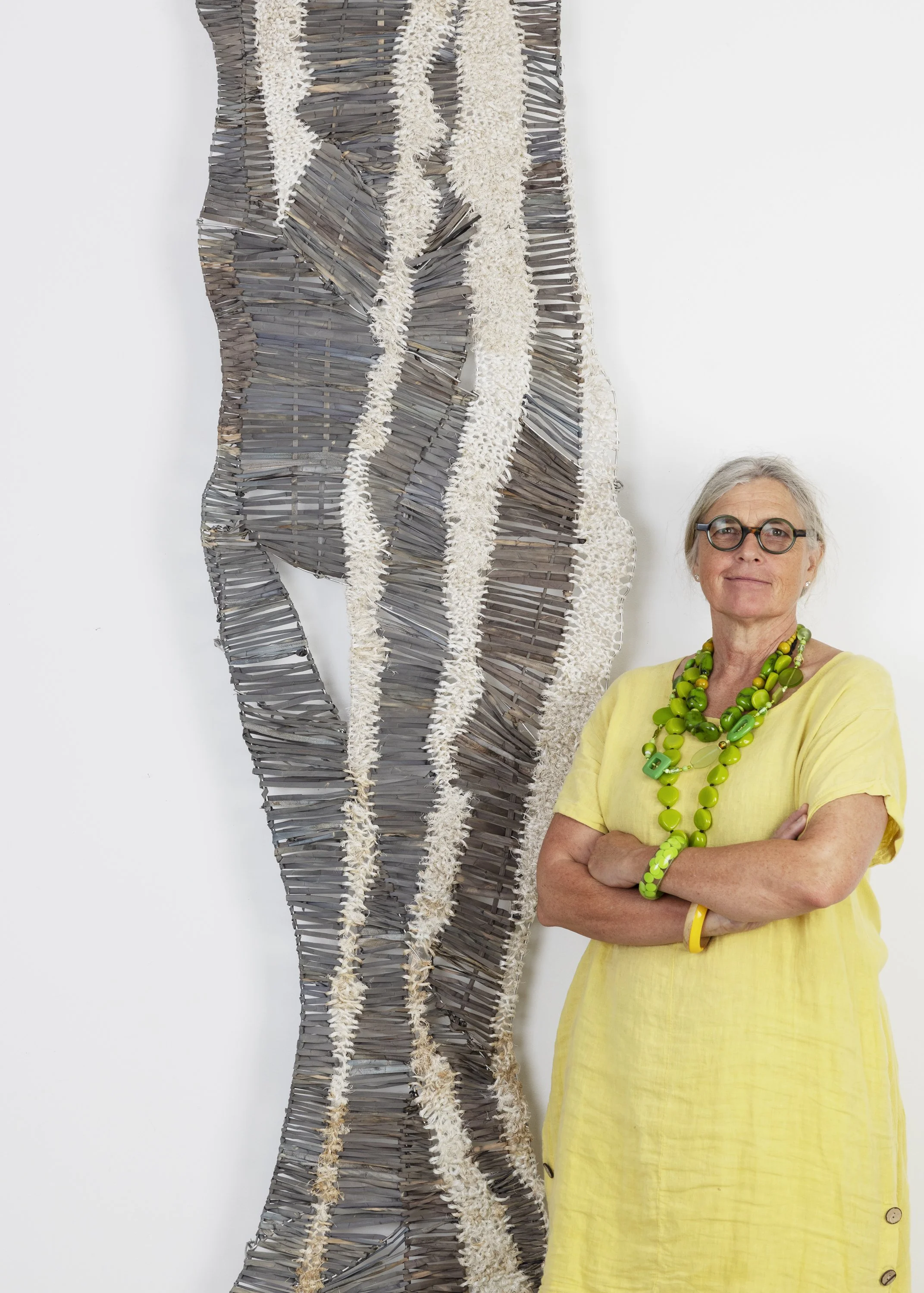 An older woman with gray hair, glasses, and a yellow dress stands with her arms crossed in front of a large, abstract sculpture made of layered wood and white textured material.