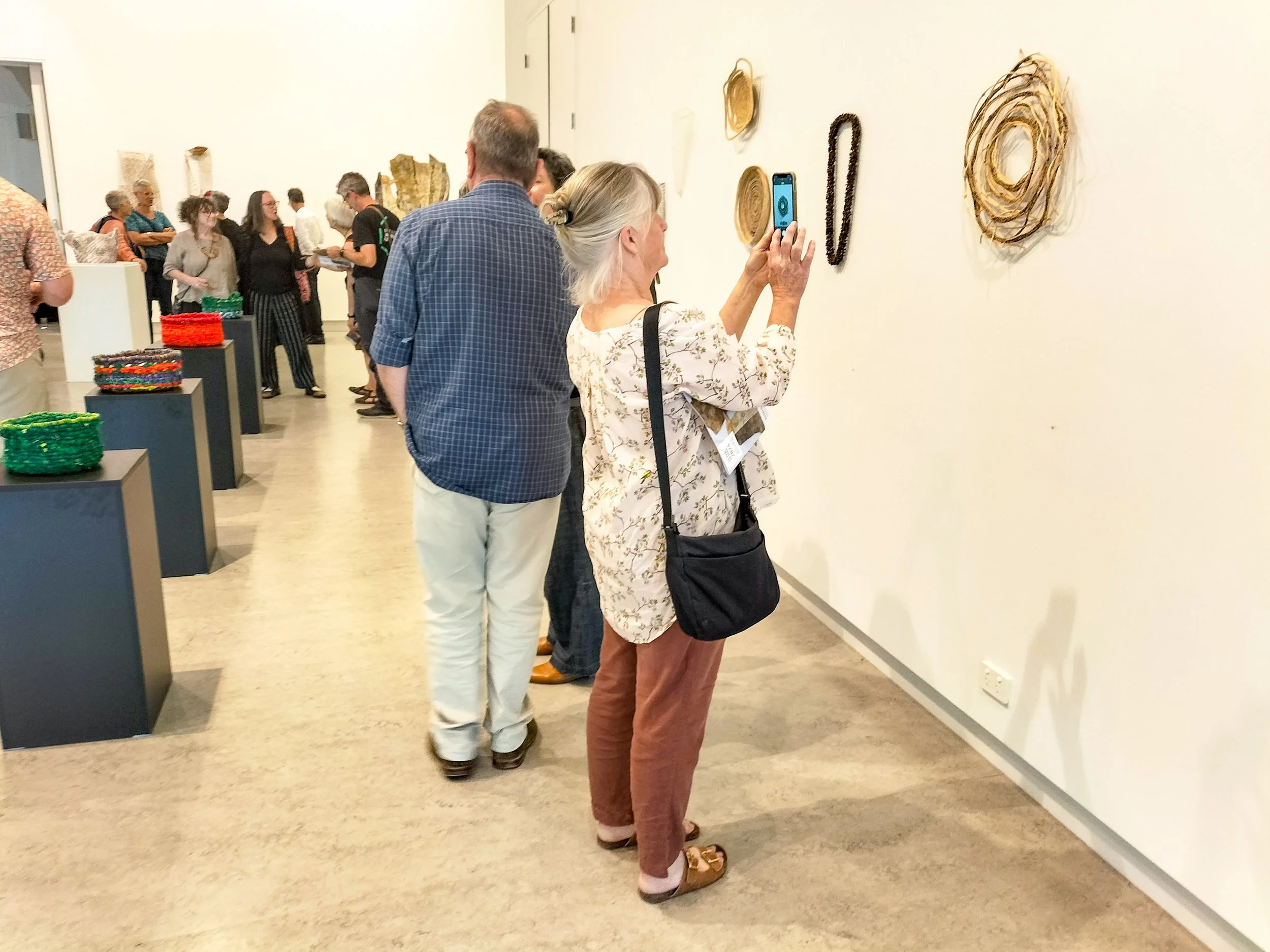 Gallery visitors viewing artwork on a white wall at an art exhibit, with colorful woven baskets on display tables nearby.