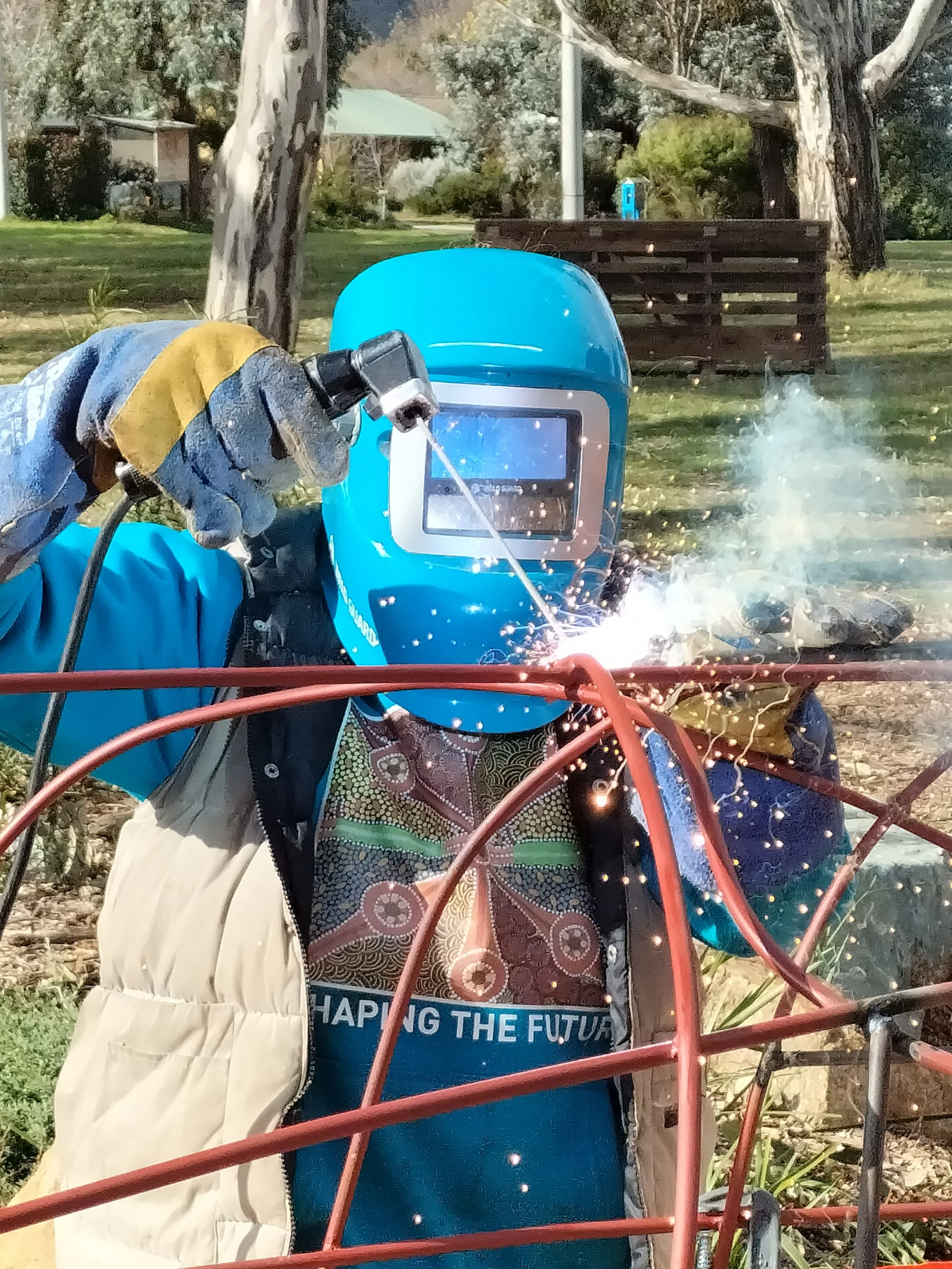 A person wearing a protective welding mask, gloves, and a blue jacket, welding metal outdoors with sparks flying.