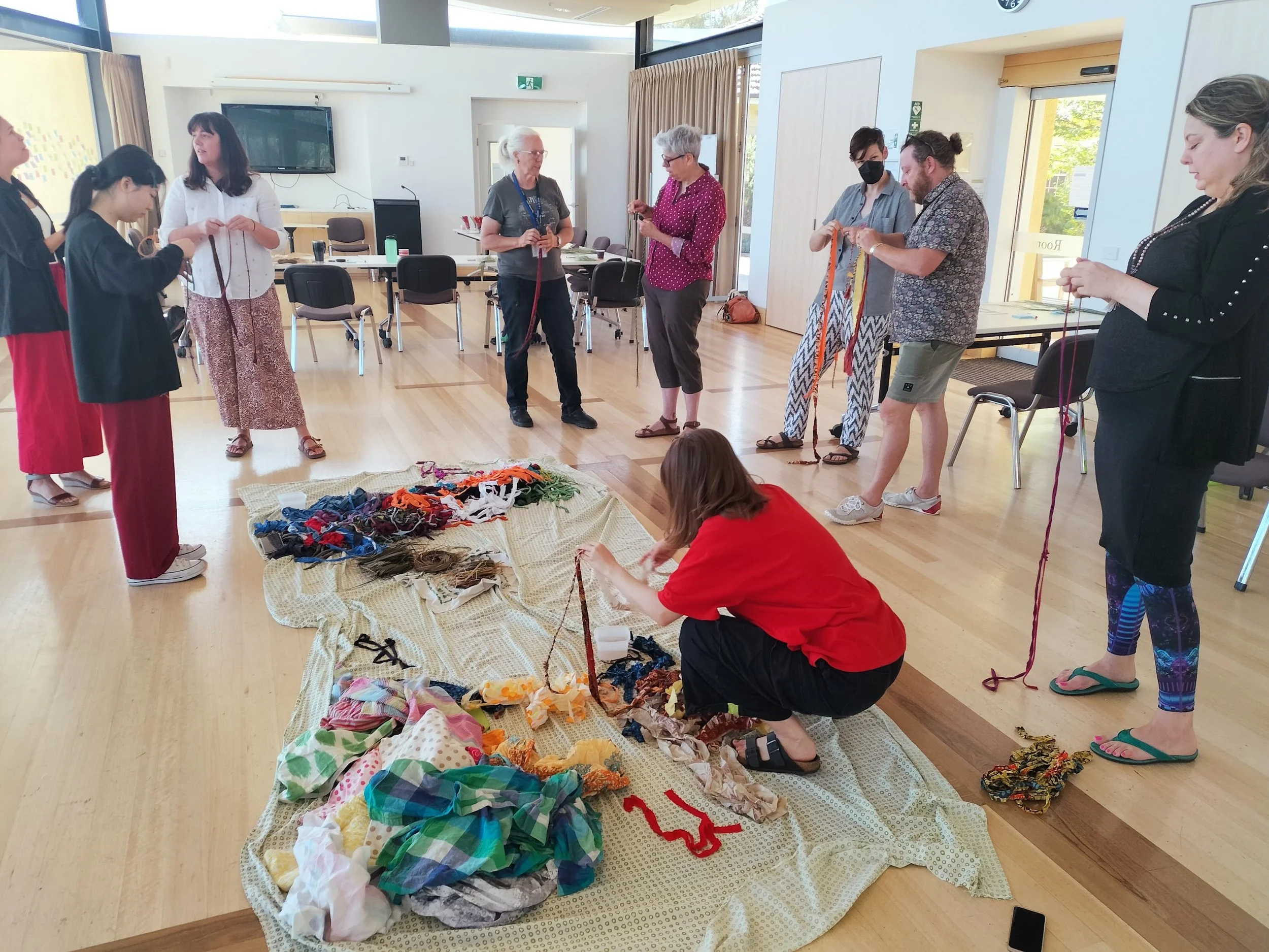 A group of seven women and one child's head are gathered in a bright room with wooden floors, participating in a textile craft activity. Some are standing, others are kneeling or crouching, working with various colorful fabric scraps and yarns spread on a large cloth on the floor. They appear focused and engaged in their craft project.