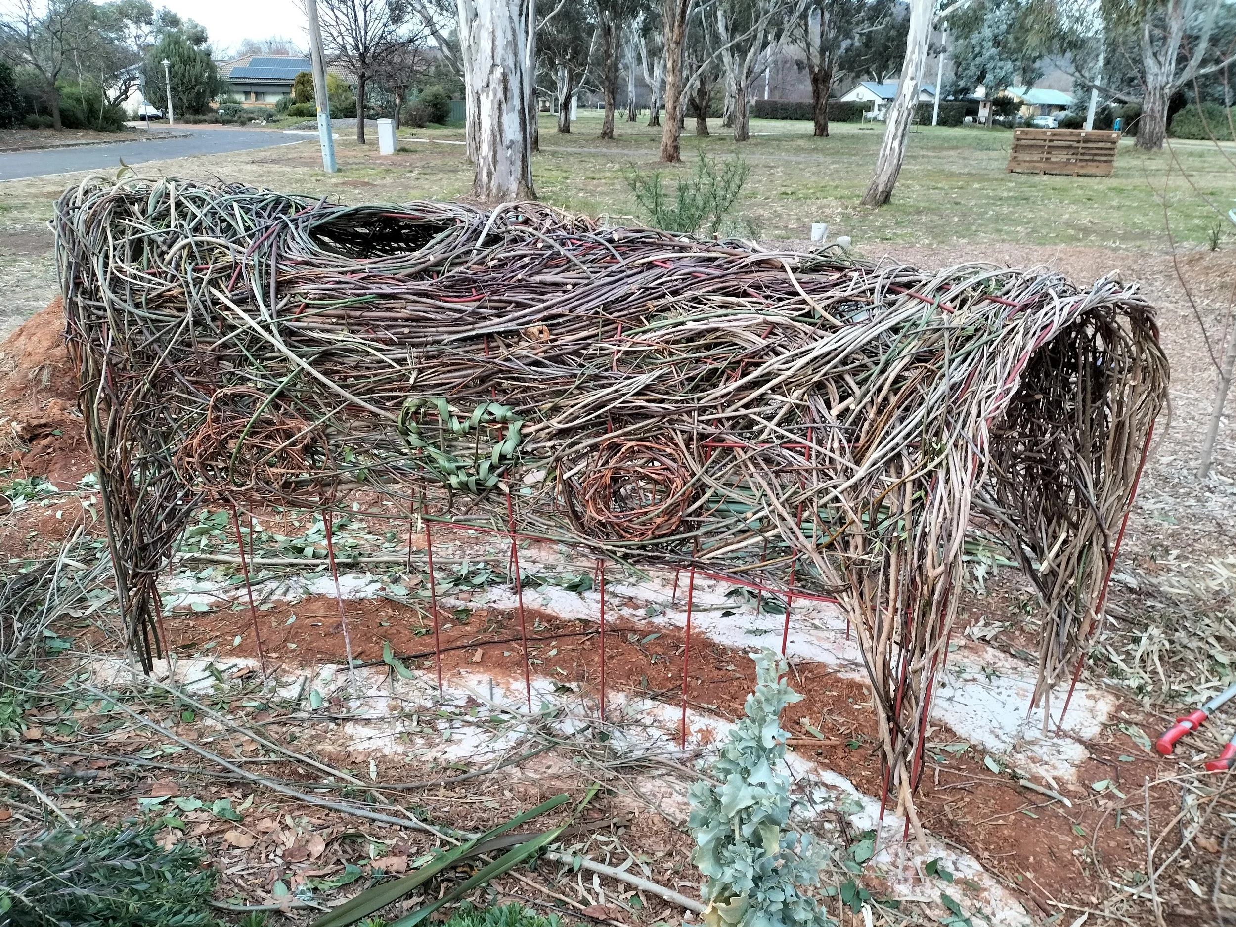 A large bird's nest inspired by a bower bird made of intertwined twigs and branches is situated on the ground in a park with trees, grass, and residential houses in the background.